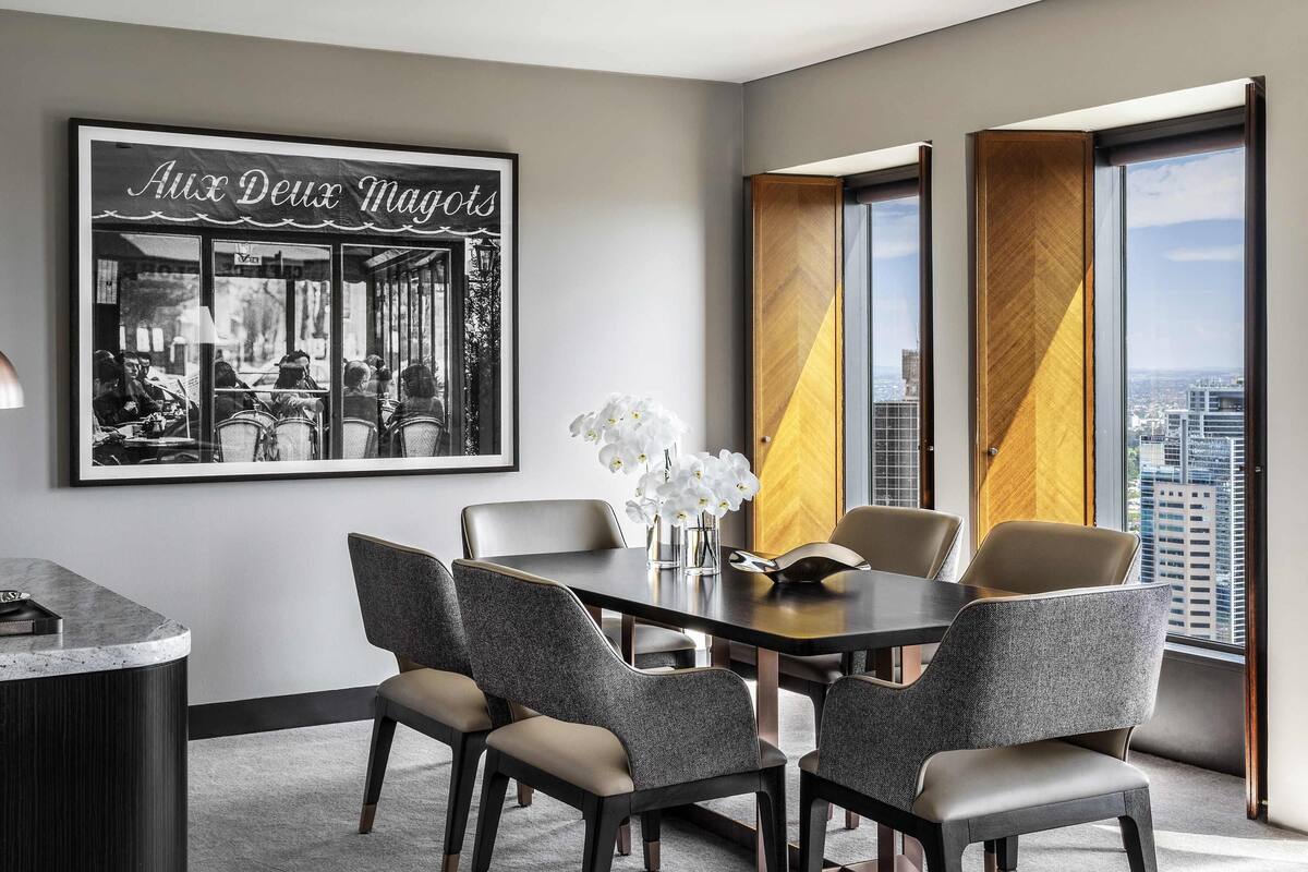 Una habitación de hotel moderna con comedor que incluye una mesa redonda y cuatro sillas, una fotografía en blanco y negro enmarcada en la pared, y ventanas grandes con cortinas.