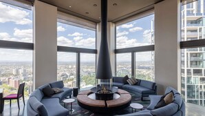 Interior de um edifício alto com área de estar circular em torno de uma lareira central, grandes janelas com vista para a cidade e design moderno.