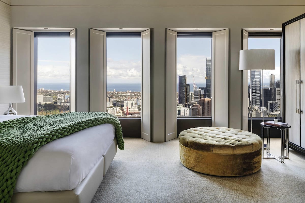 A bedroom with a bed, a green blanket, a round ottoman, and large windows with a city view.