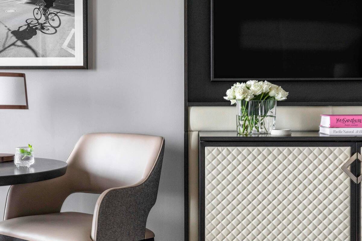 Una habitación de hotel moderna con un televisor de pantalla plana grande, un sillón cómodo, una mesita redonda con un vaso y un florero con flores blancas.