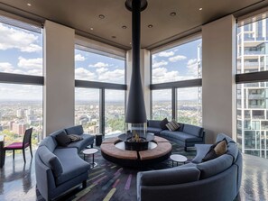 Relax in a spacious room with a circular seating area and fire pit, surrounded by sofas and armchairs, offering expansive city views.