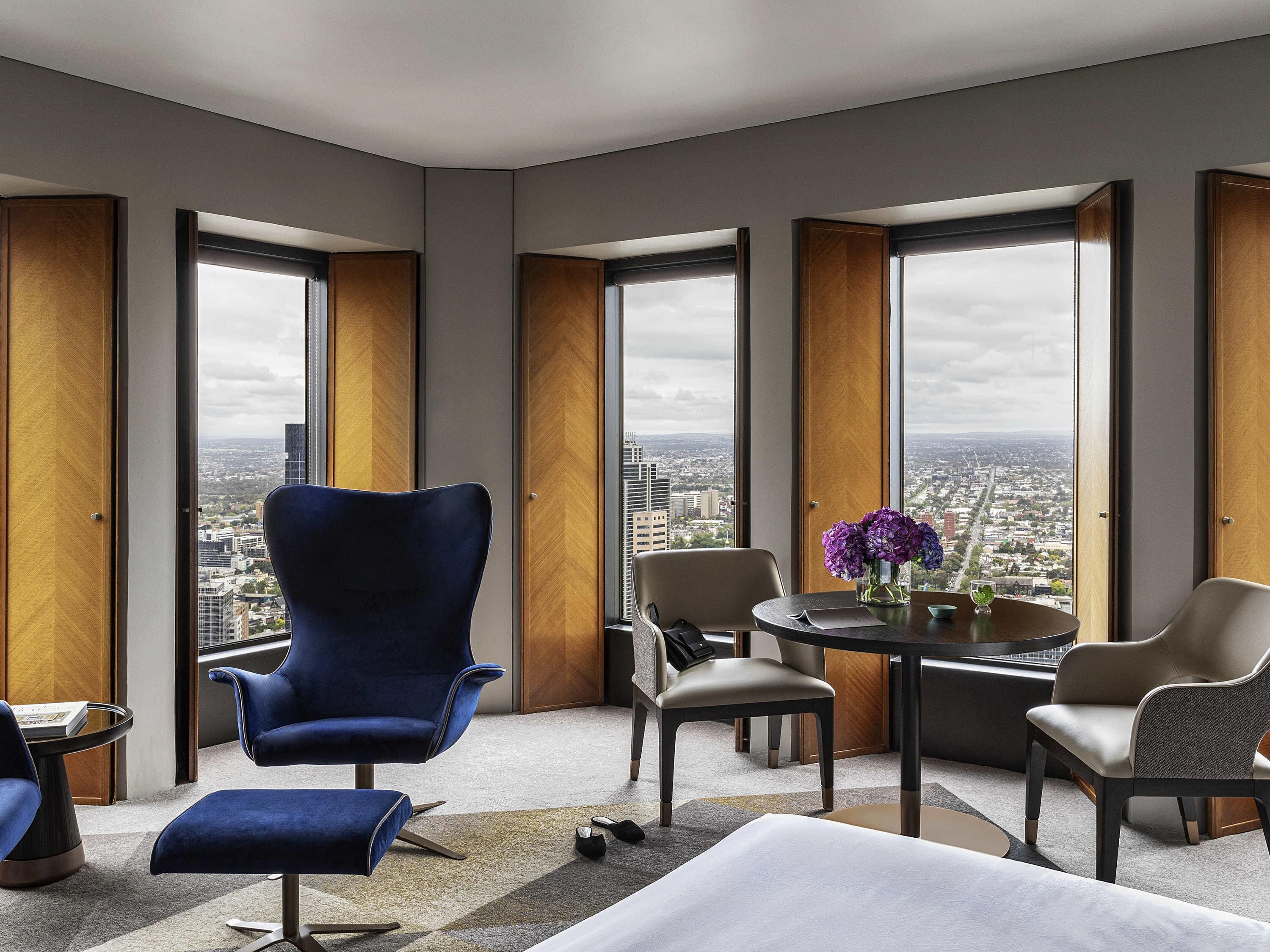 A modern hotel room with a large city view, a blue armchair, a round table with a vase of flowers, and a sofa.