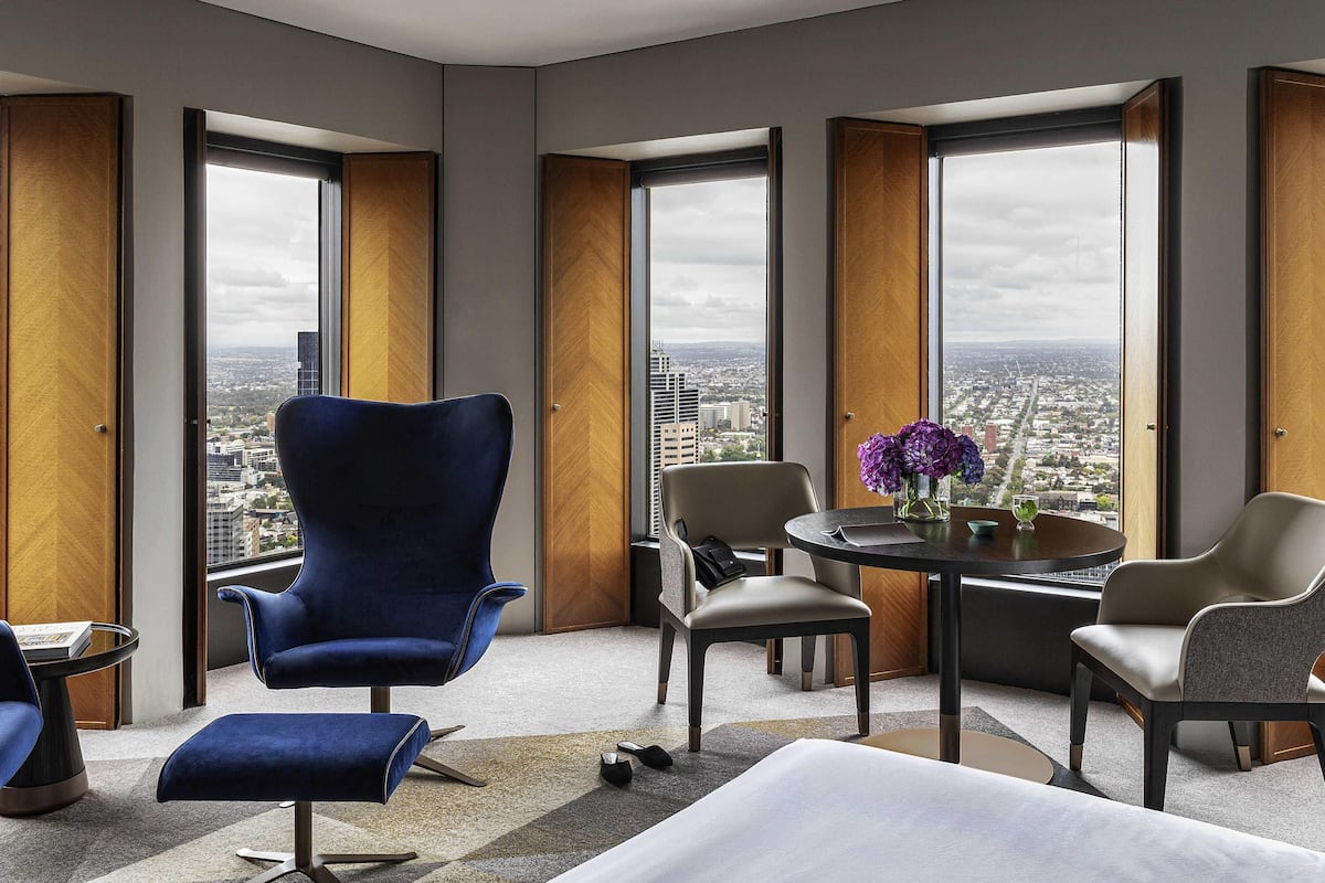 A modern hotel room with a large city view, a blue armchair, a round table with a vase of flowers, and a sofa.