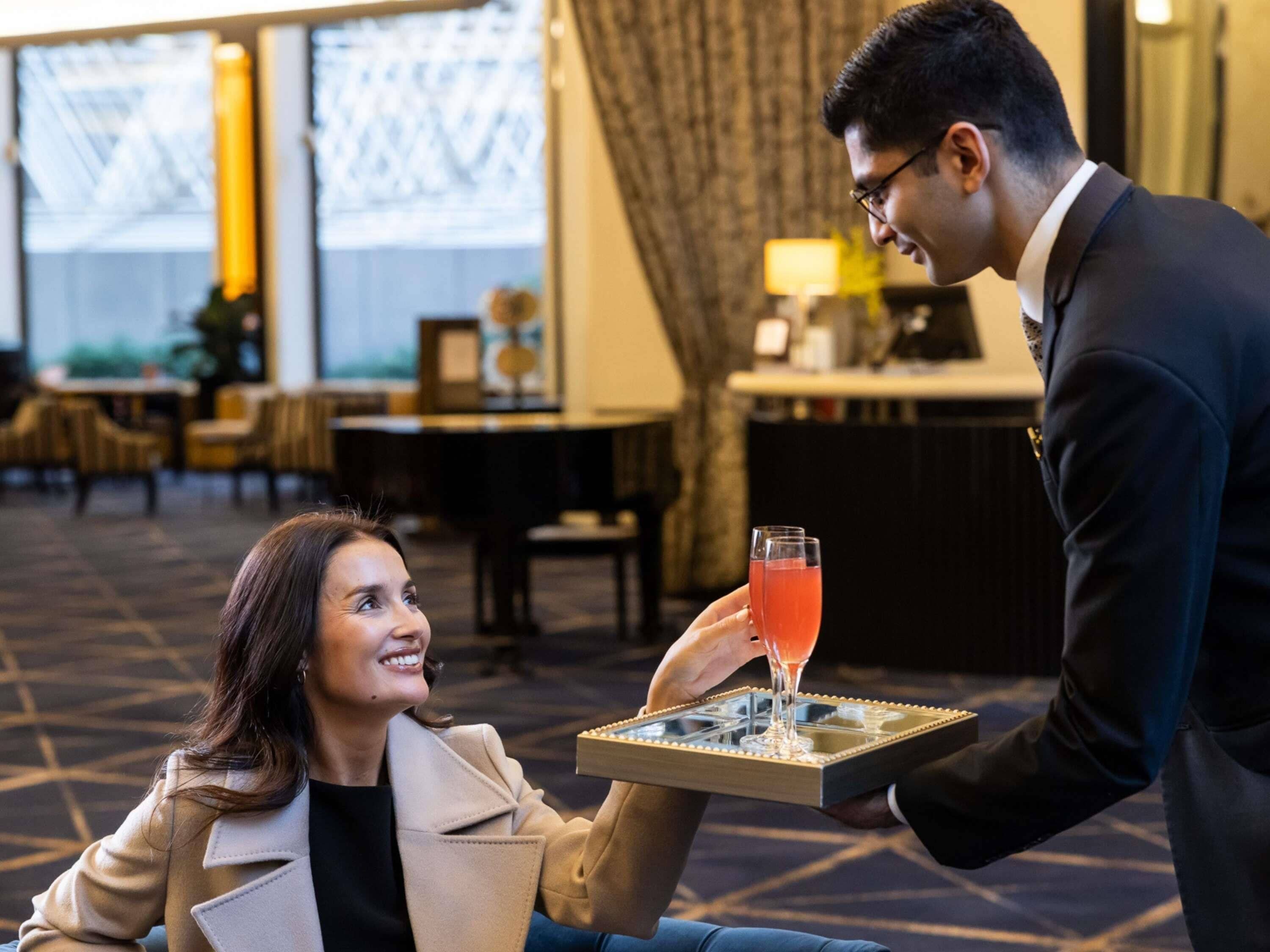 A person in a suit serving a drink to a seated individual in a hotel setting with a bar and seating area in the background.