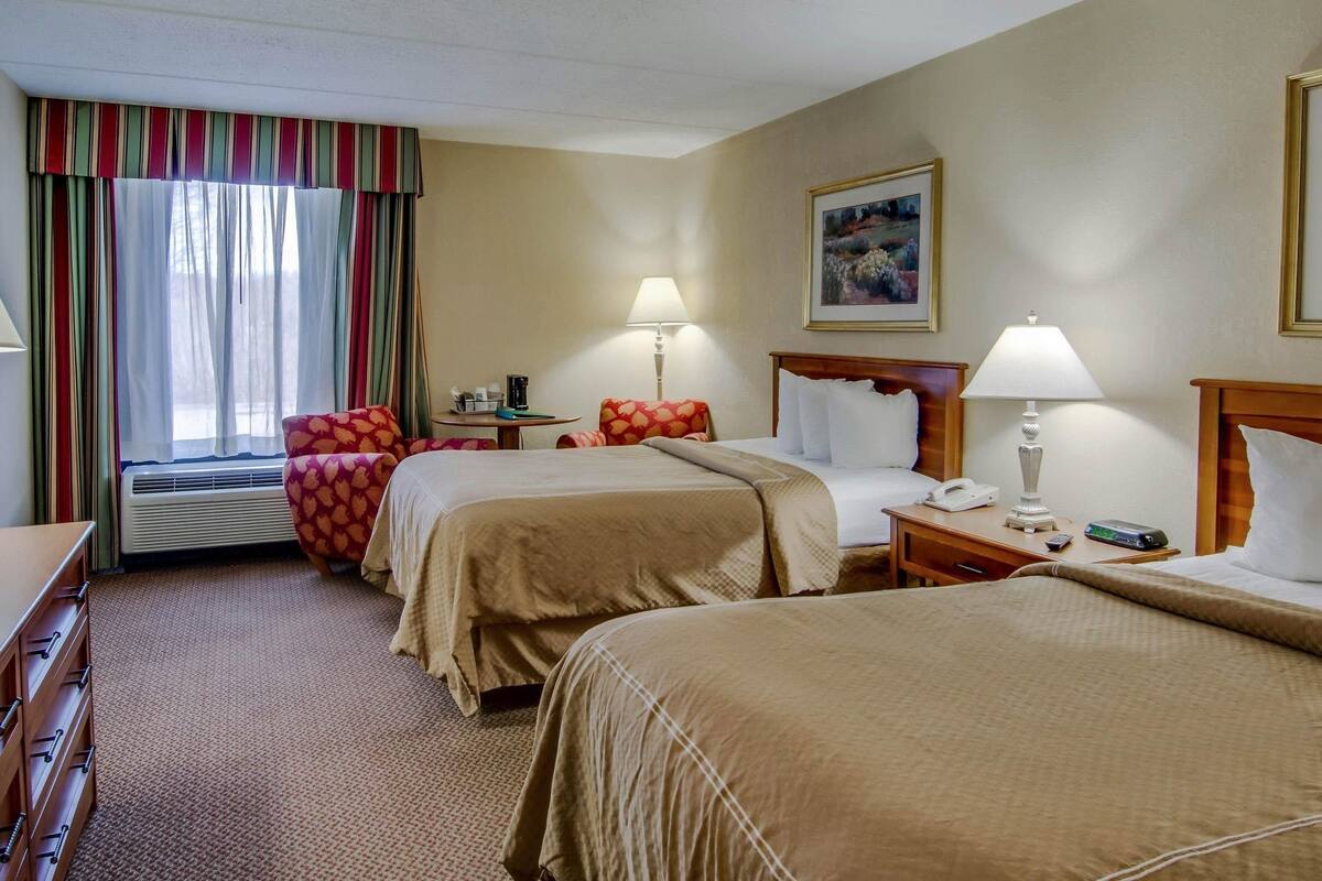 A hotel room with two beds, a desk, a chair, a table with a lamp, a painting on the wall, and a window with curtains.