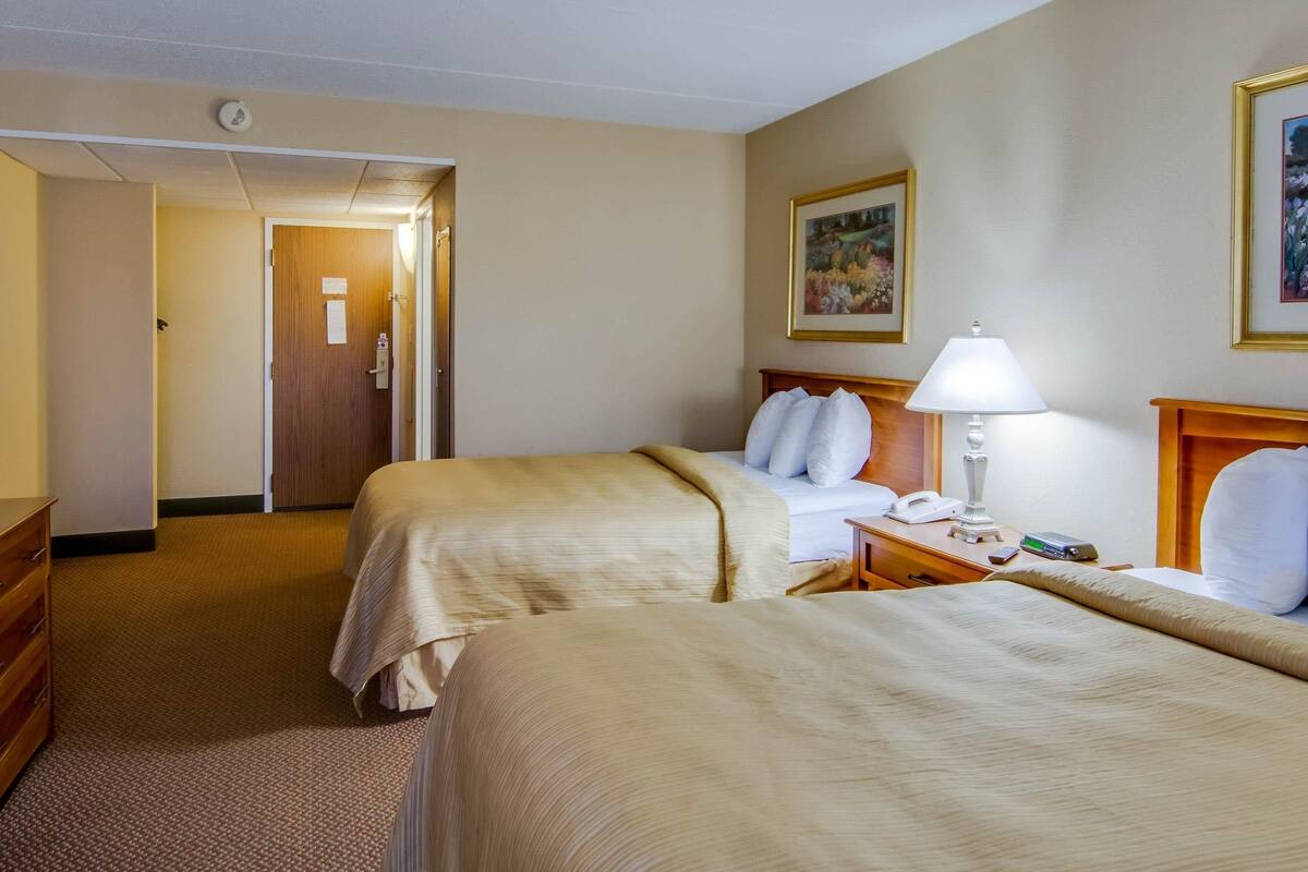 A hotel room with two beds, a wooden headboard, a bedside table with a lamp, a phone, and a painting on the wall.