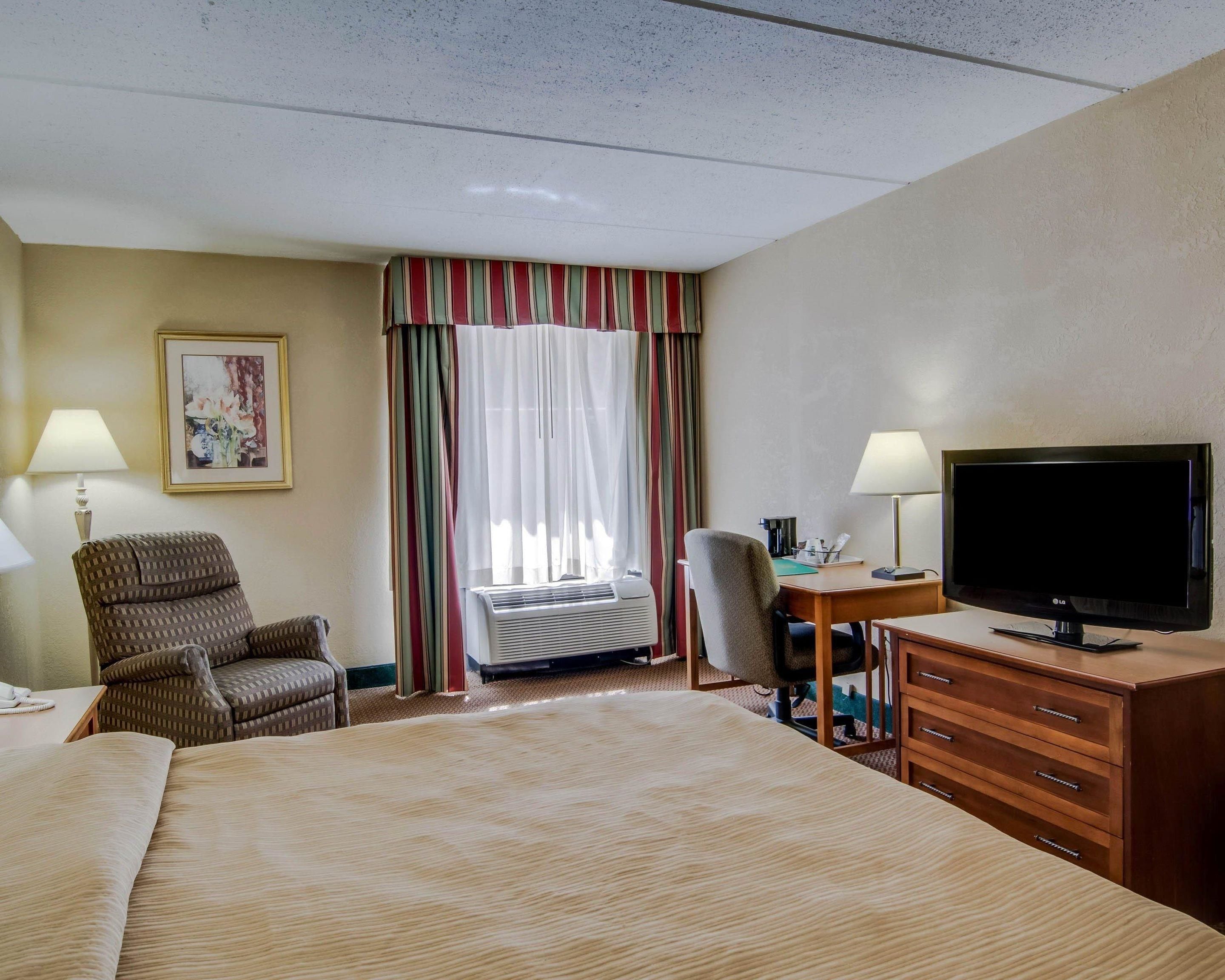 A hotel room with a bed, a desk with a TV, a chair, a desk lamp, and an air conditioning unit.