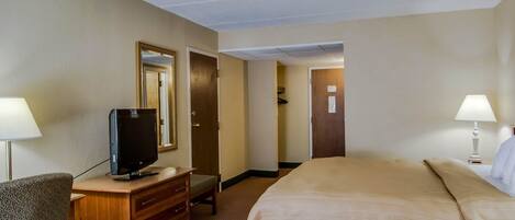 Een hotelkamer met een bed, een bureau met een televisie, een kast, twee lampen en een deur naar een andere kamer.