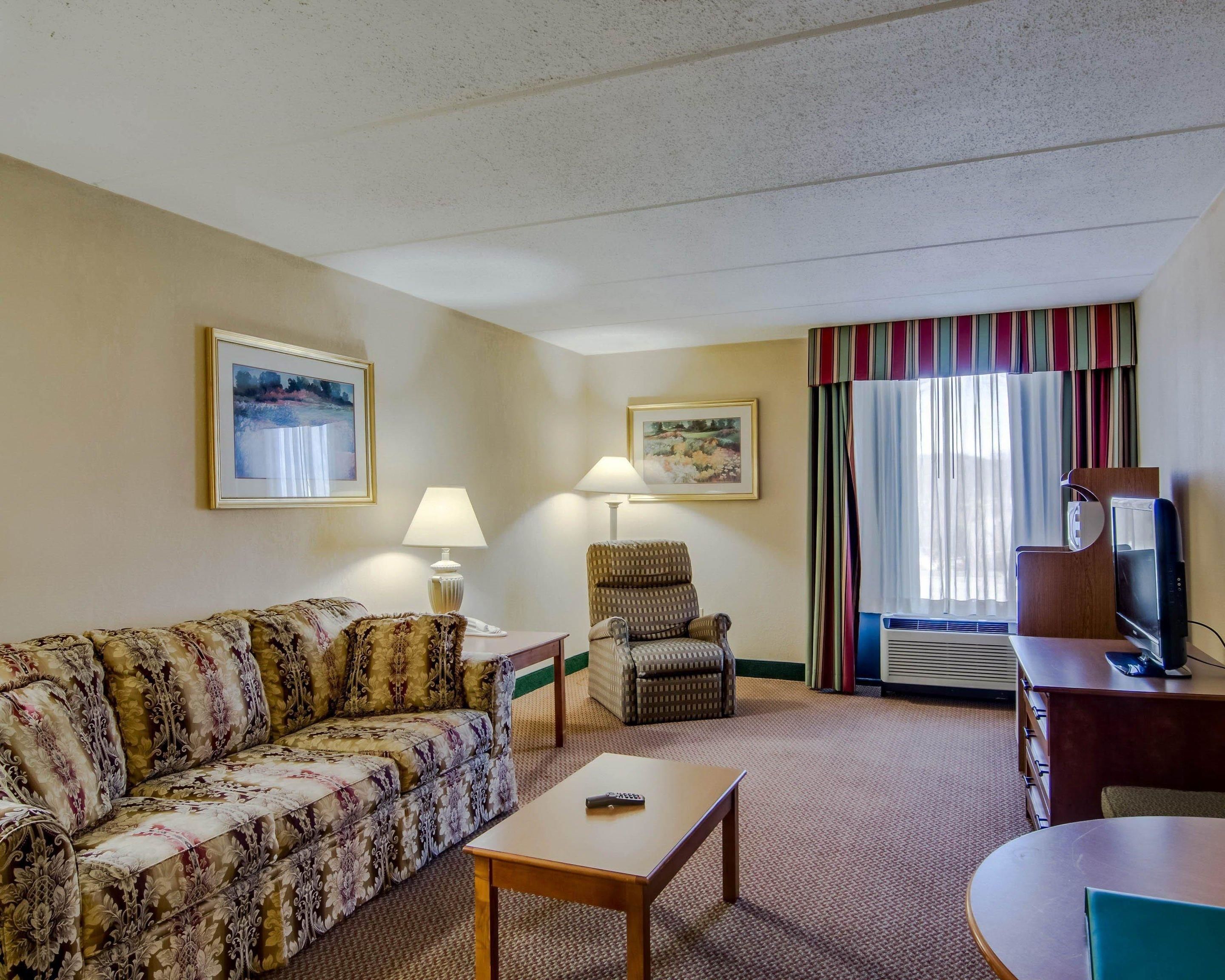 A hotel room with a sofa, armchair, table, and a TV stand with a remote control.