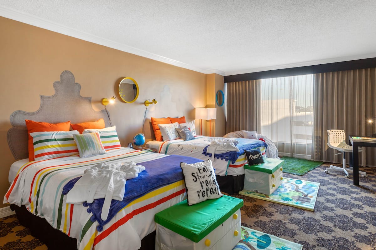 A hotel room with two beds, a desk, a chair, and a large window with curtains.