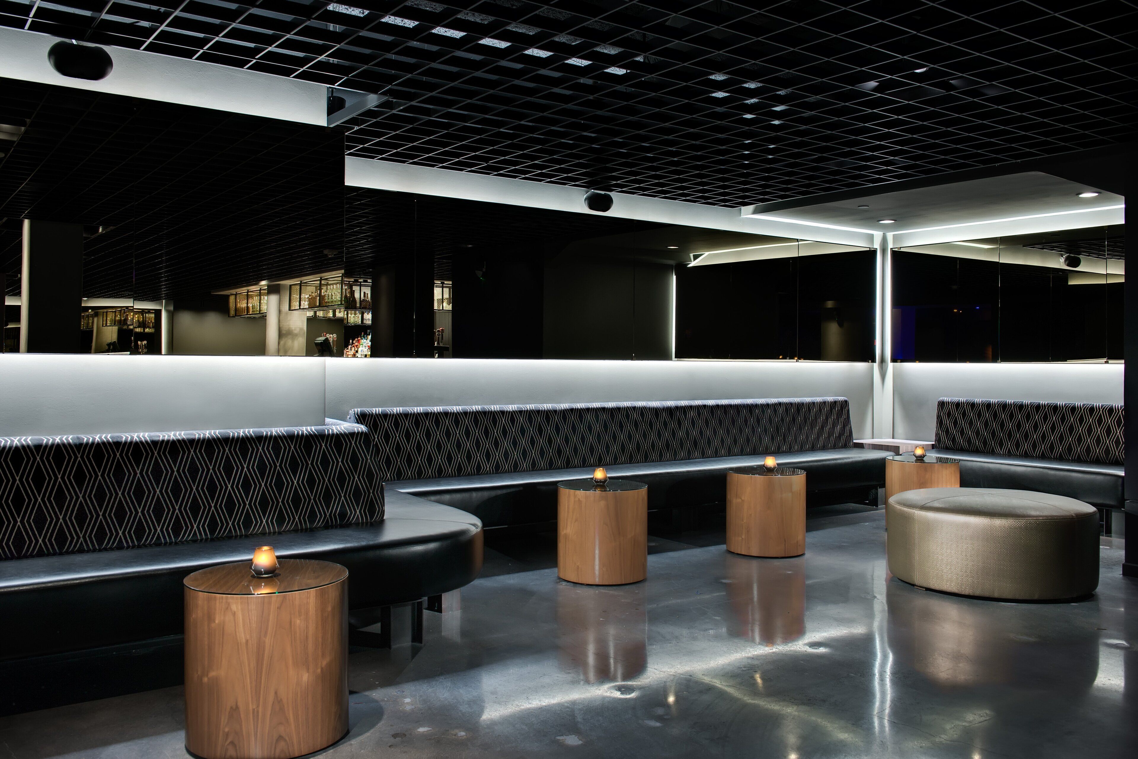 A modern interior with a seating area featuring black cushioned benches and wooden stools, a polished concrete floor, and a grid-patterned ceiling.