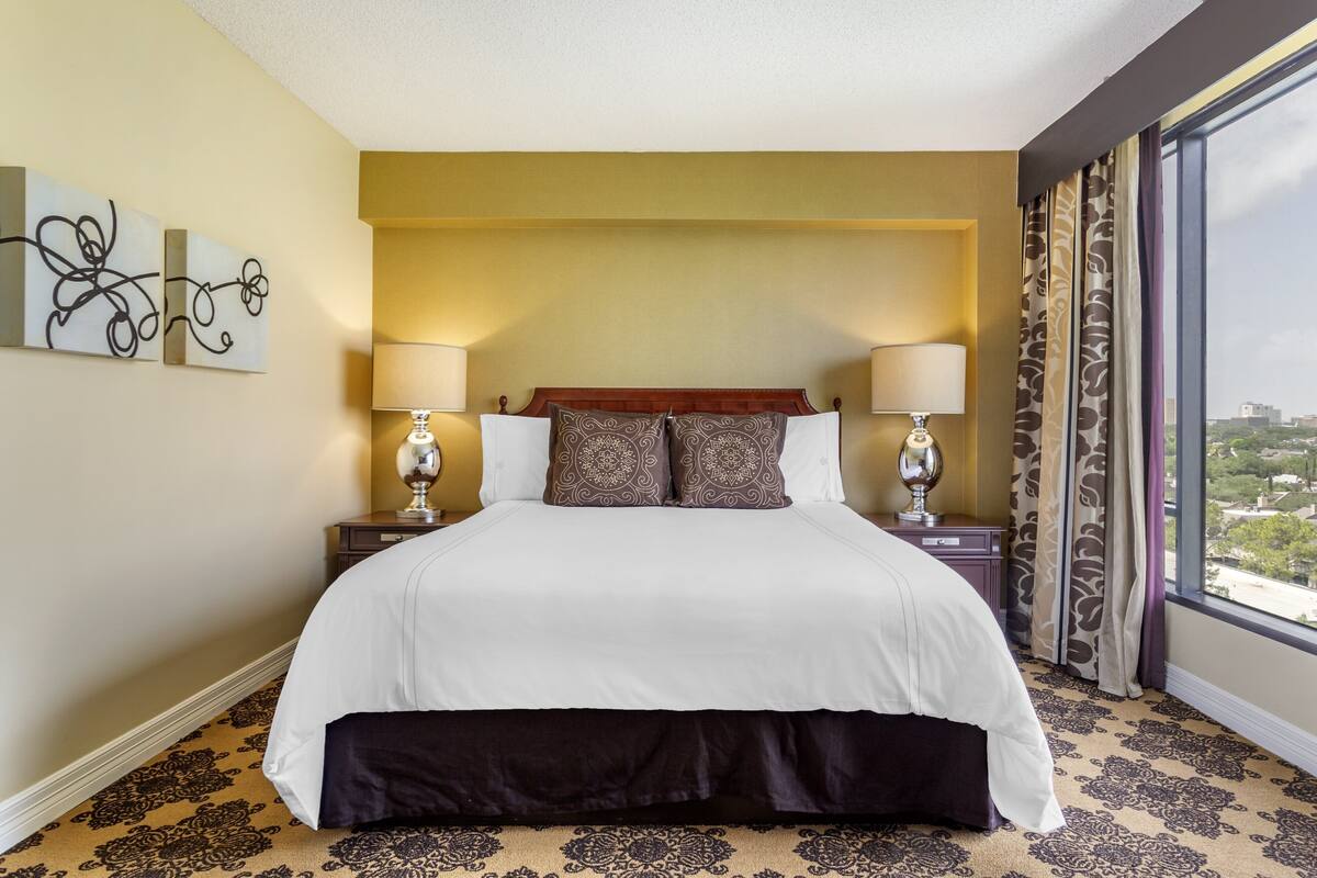 A hotel room with a large bed, two bedside tables with lamps, a view of the city through a large window, and a patterned carpet.