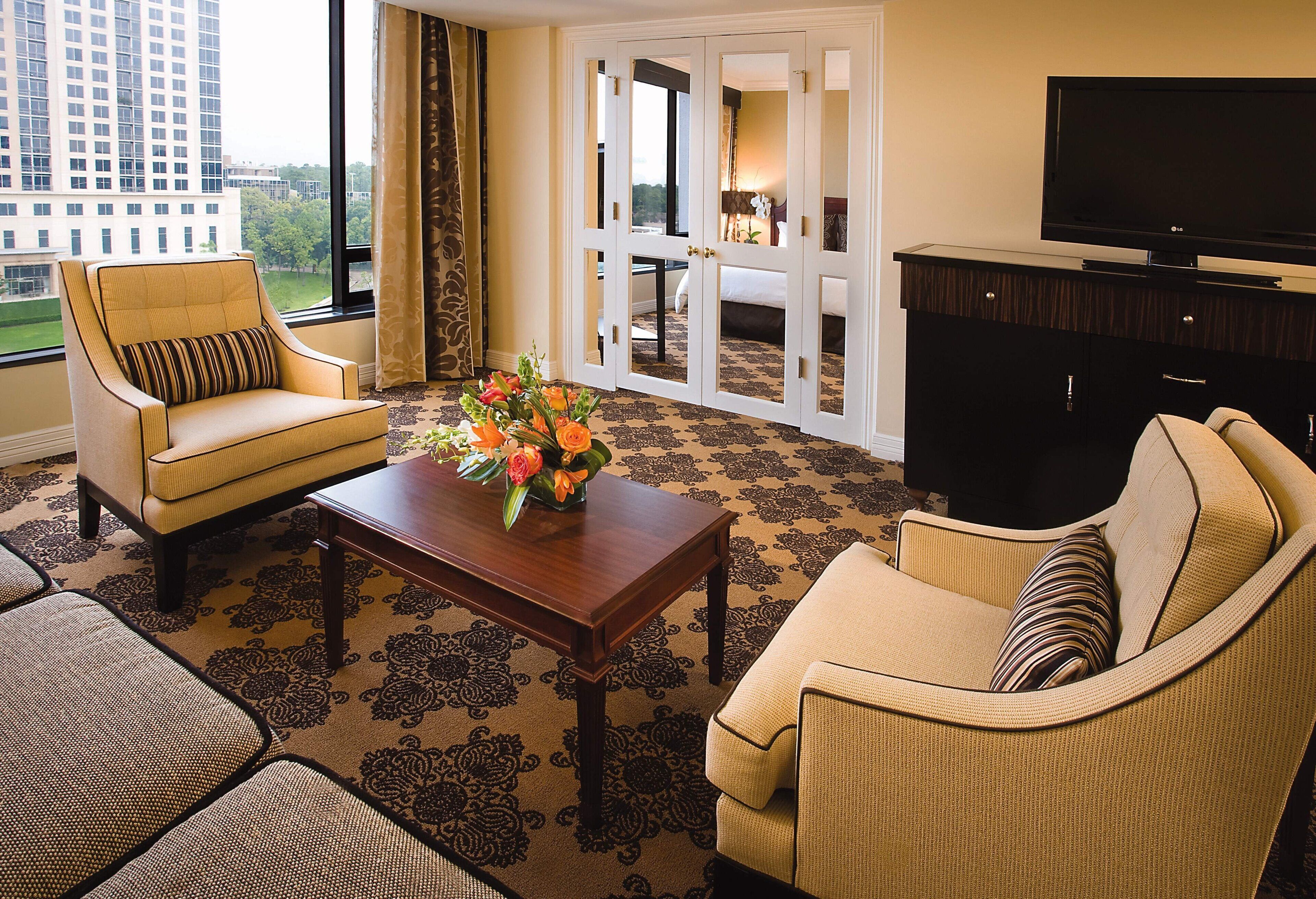 A hotel room with a large window, a sofa, a chair, a coffee table with flowers, and a TV on a cabinet.