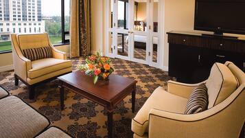 A hotel room with a large window, a sofa, a chair, a coffee table with flowers, and a TV on a cabinet.