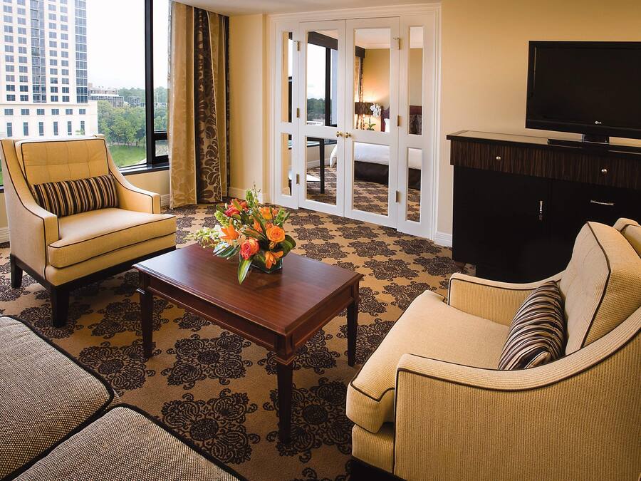 A hotel room with a large window, a sofa, a chair, a coffee table with flowers, and a TV on a cabinet.