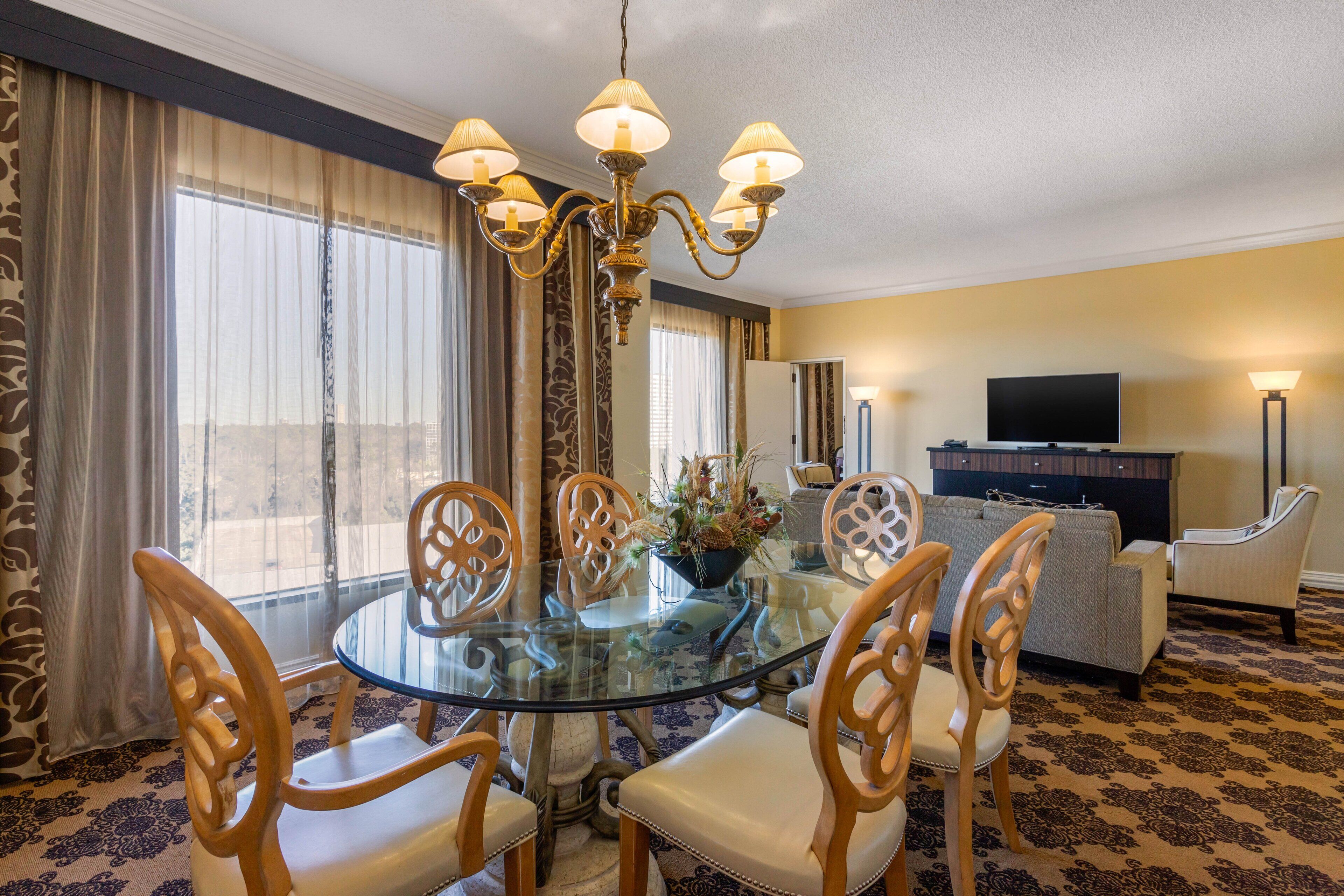 A hotel room with a round glass table, wooden chairs, a chandelier, a sofa, a television, and a large window with curtains.