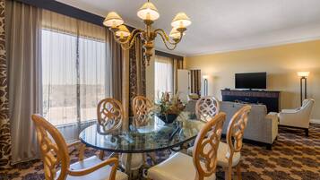 A hotel room with a round glass table, wooden chairs, a chandelier, a sofa, a television, and a large window with curtains.