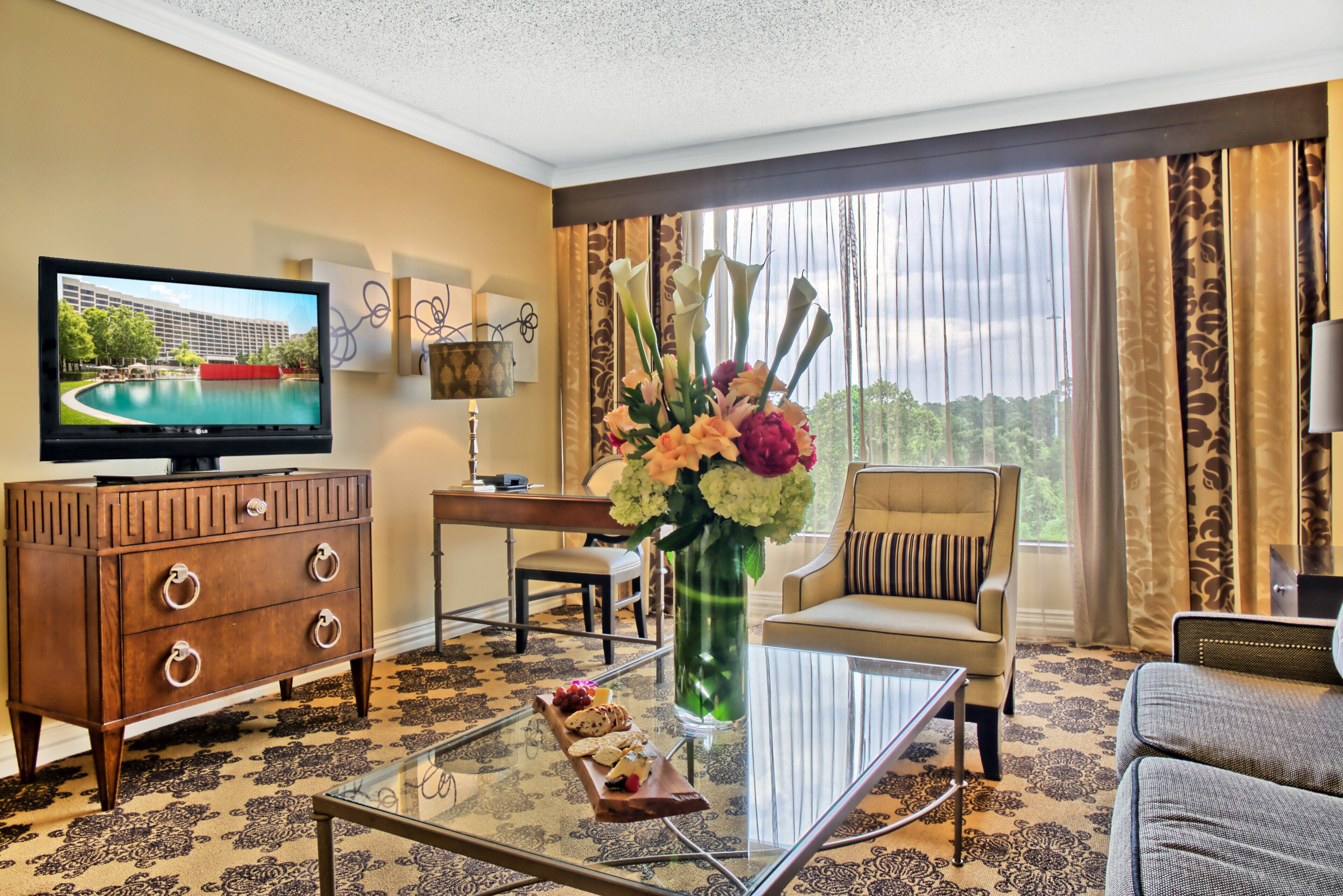 A hotel room with a television, a wooden dresser, a glass coffee table with snacks, a vase of flowers, a sofa, and a view of a building through large windows.
