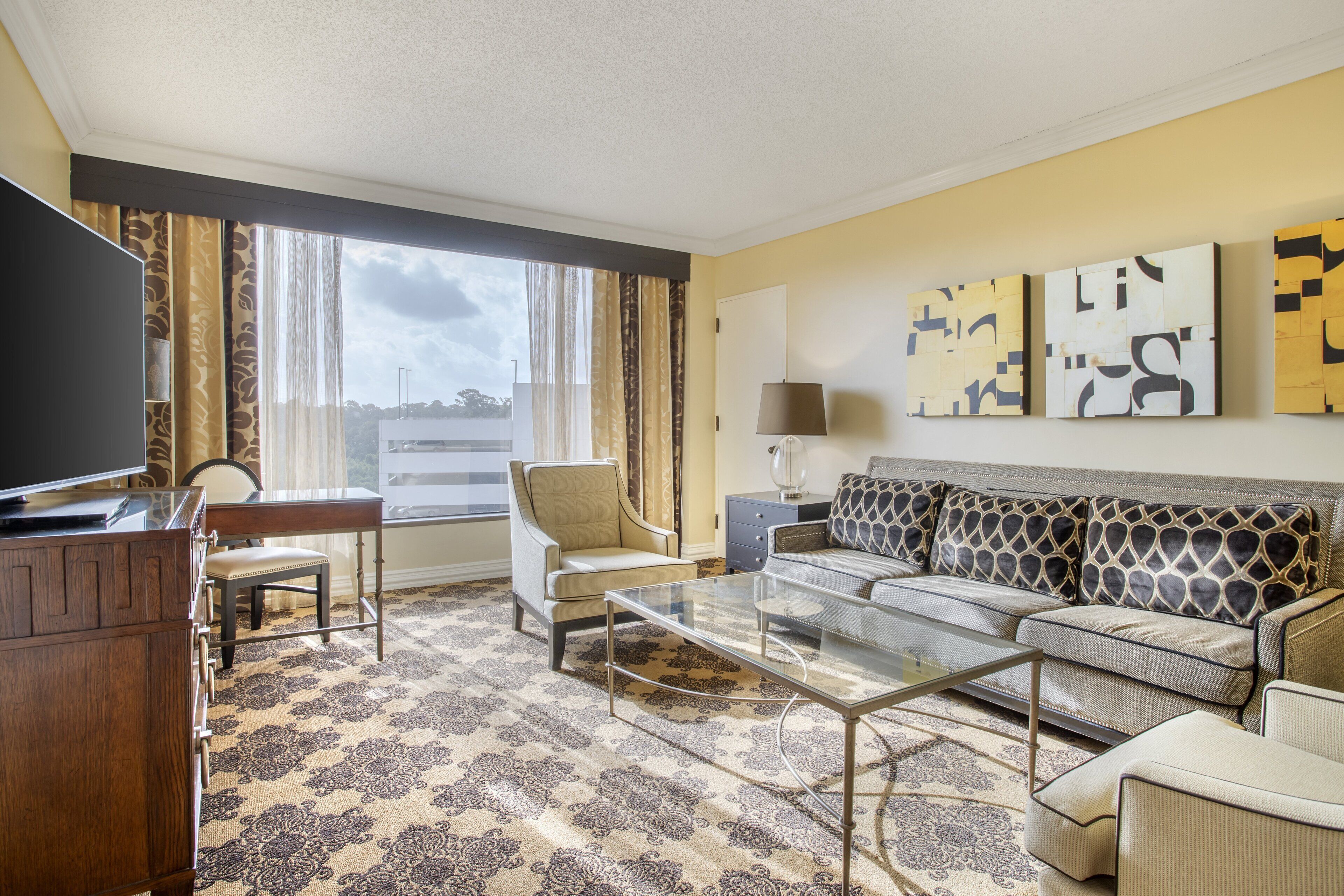 A hotel room with a sofa, armchair, glass coffee table, TV, and artwork on the wall.