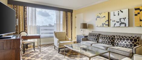 A hotel room with a sofa, armchair, glass coffee table, TV, and artwork on the wall.