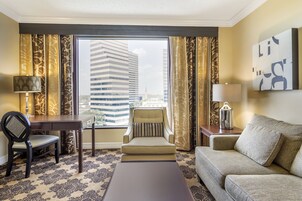 Quarto de hotel com uma grande janela, uma escrivaninha, uma cadeira, um sofá e uma luminária.