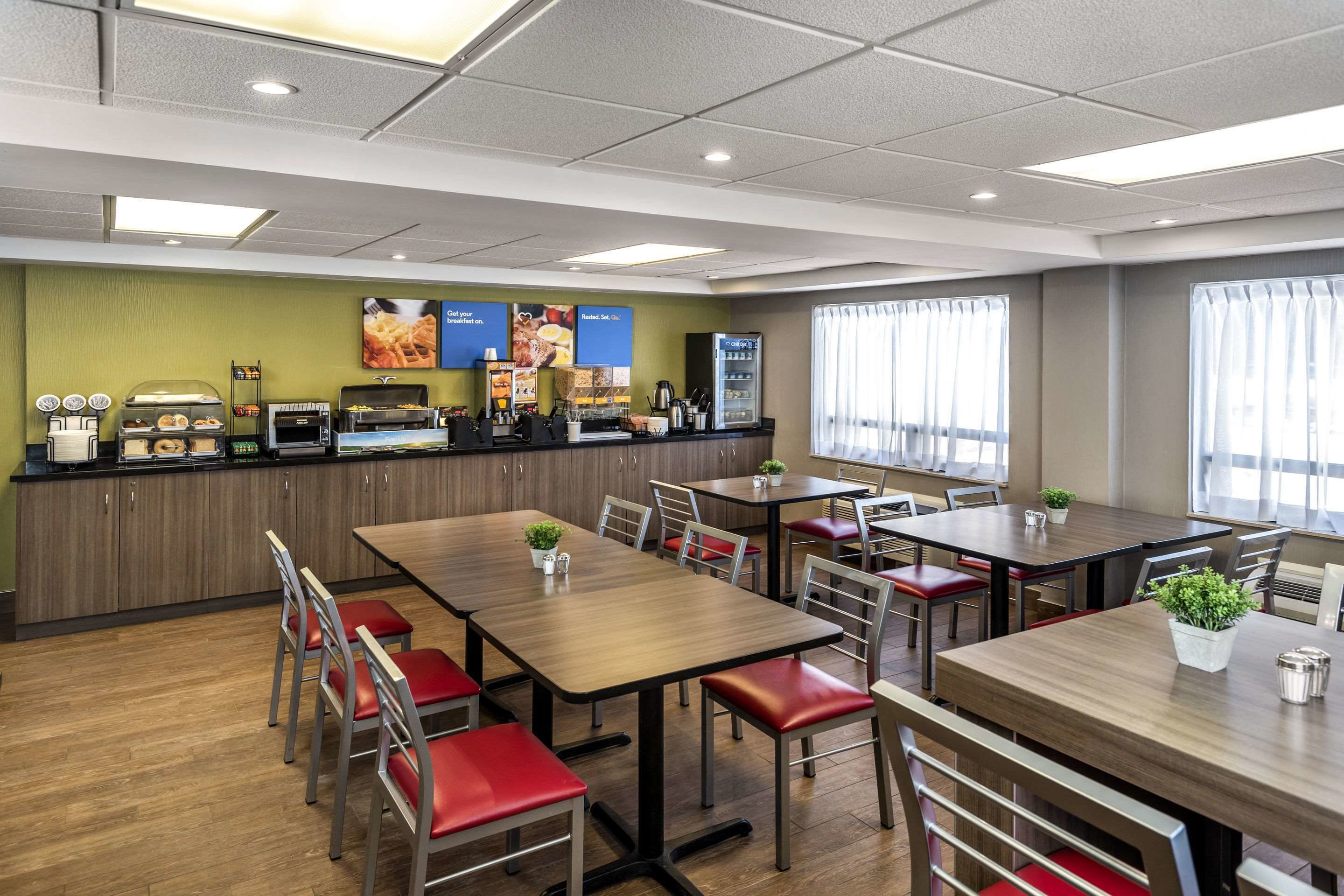 A modern restaurant with wooden tables and red cushioned chairs, a counter with food items, and a view of the city through large windows.