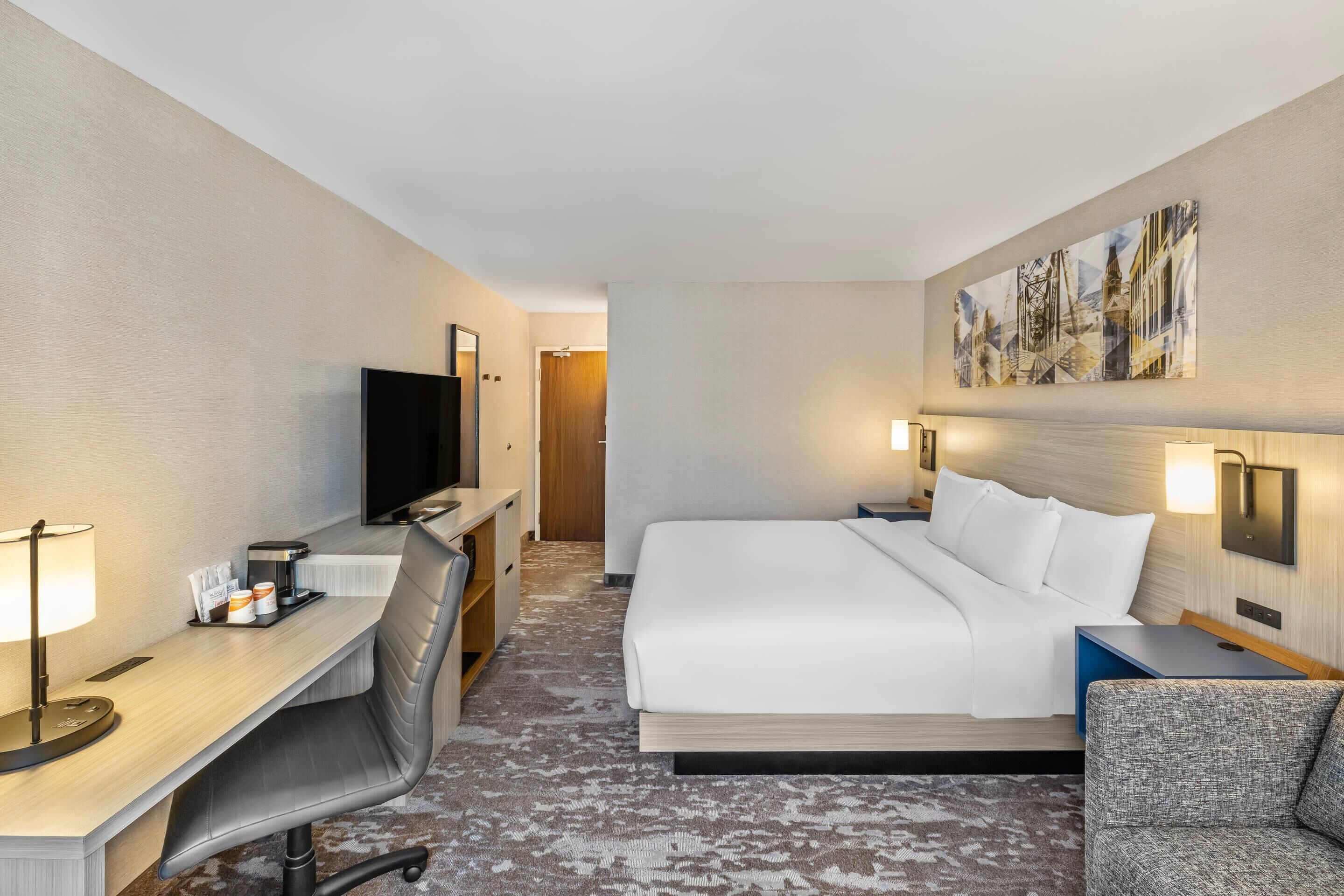 A hotel room with a bed, a desk with a chair, a television, and a painting on the wall.