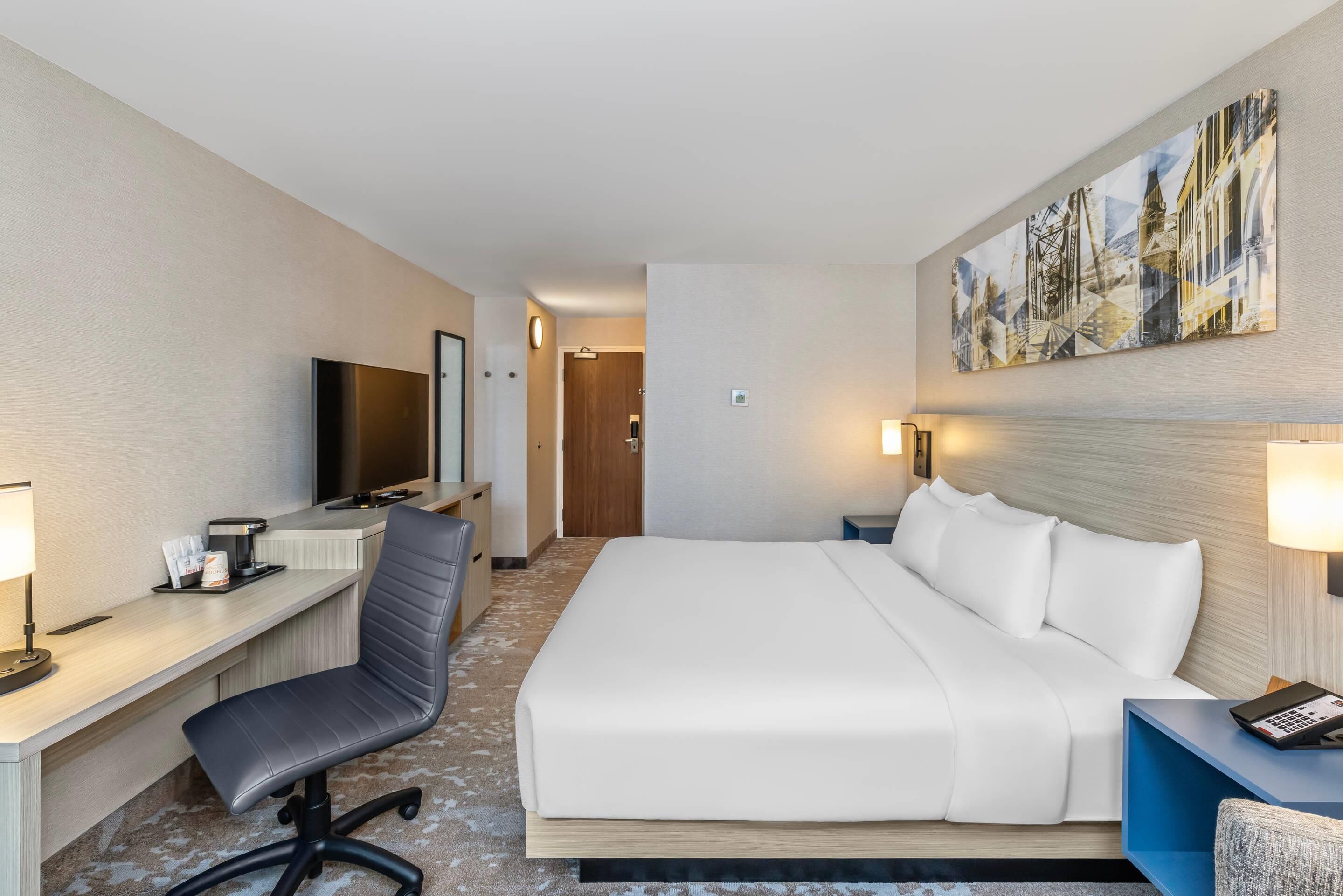 A hotel room with a bed, desk, chair, TV, and artwork on the wall.