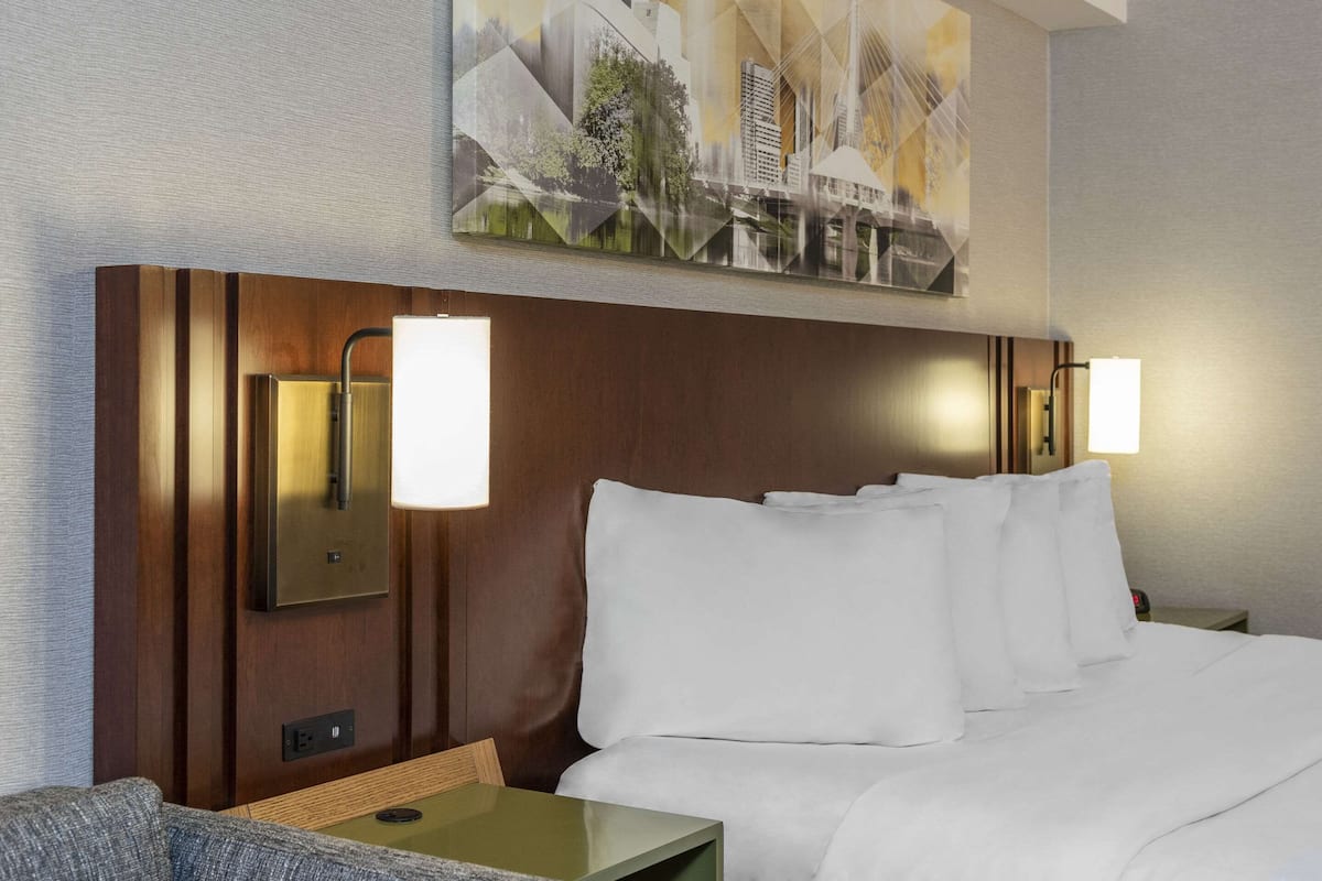 A hotel room with a wooden headboard, two bedside lamps, a bed with white linens, and a painting above the headboard.