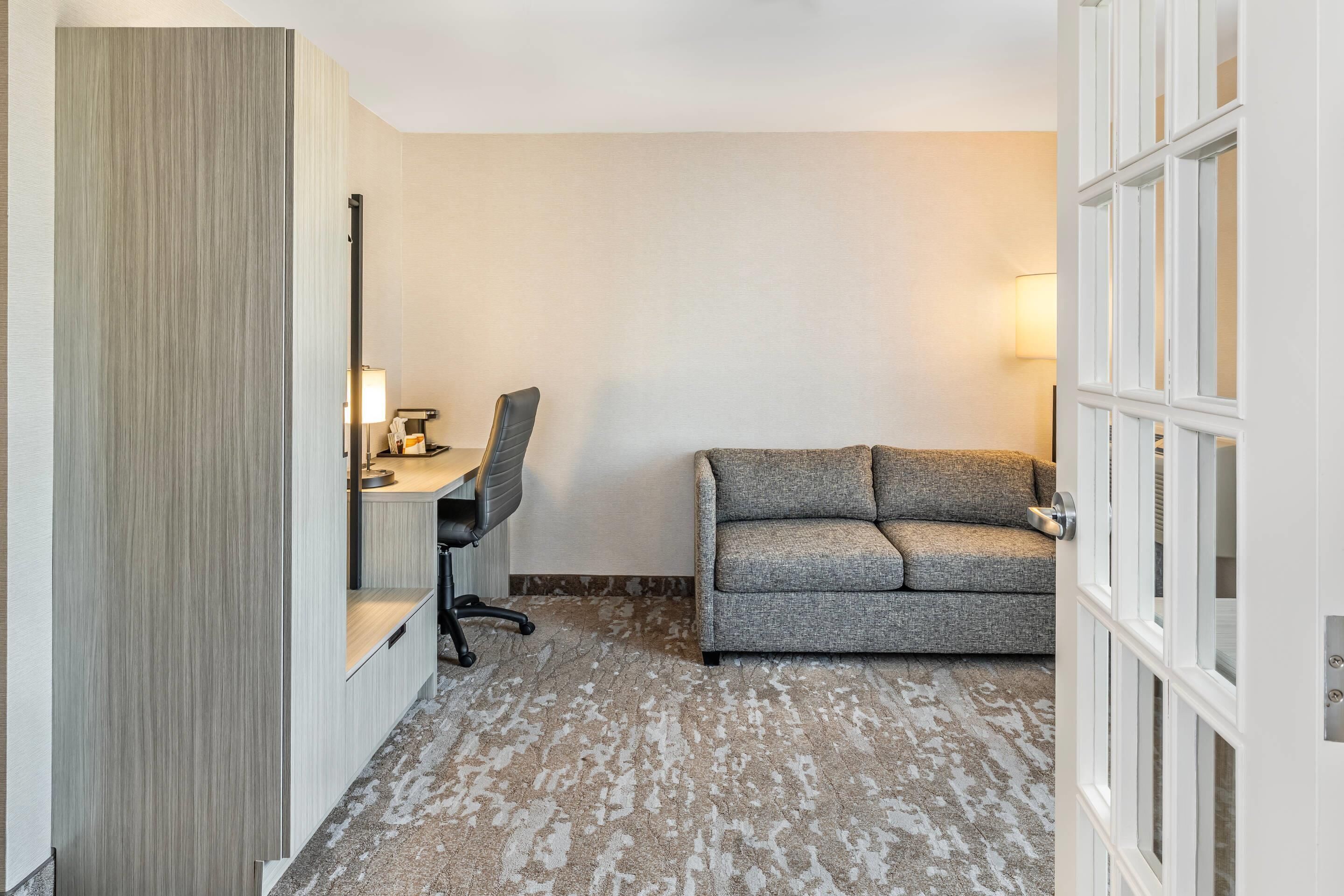 A hotel room with a sofa, a desk with a chair, a lamp, and a large window.