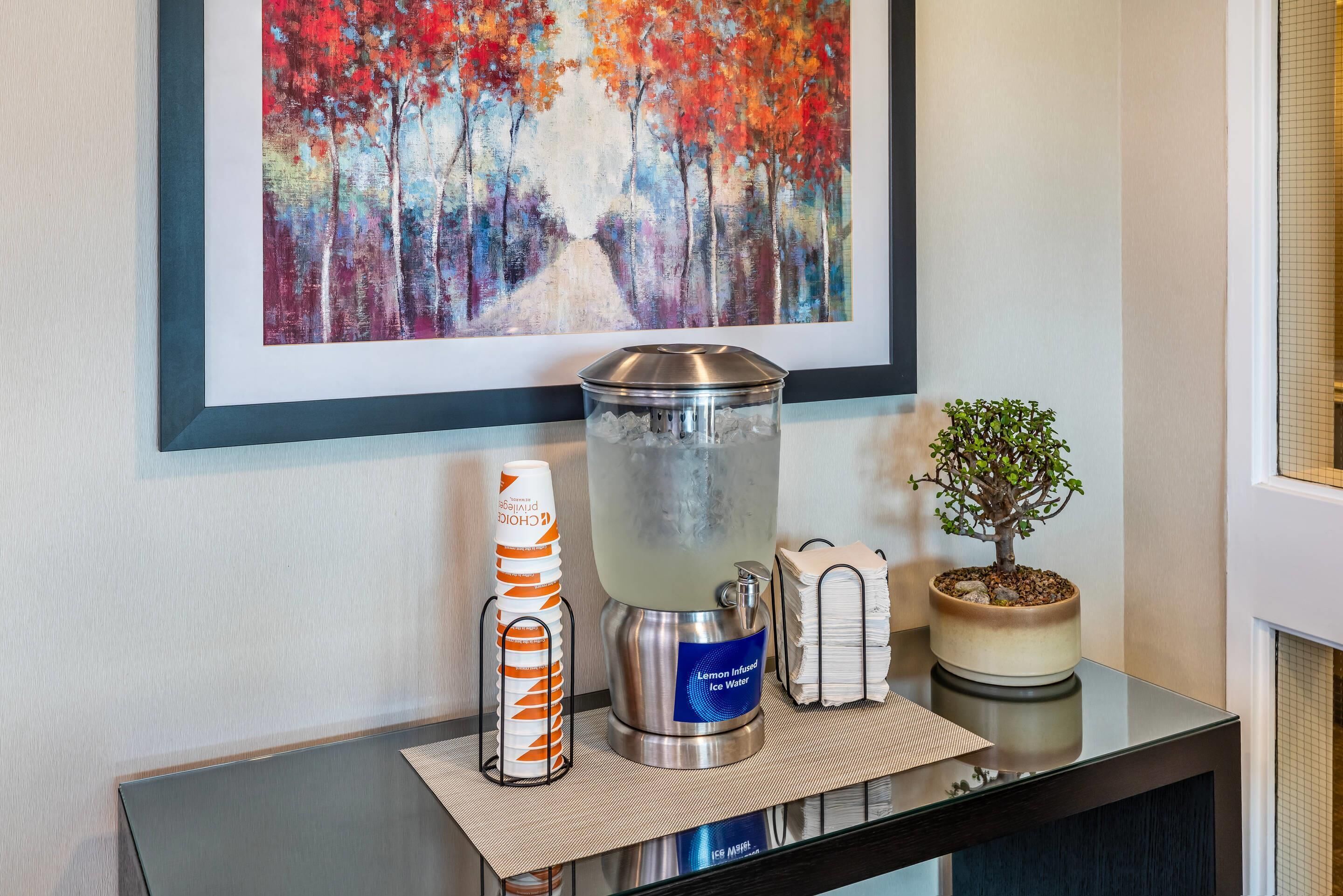 A water dispenser, a stack of napkins, a small potted plant, and a framed painting on a table.