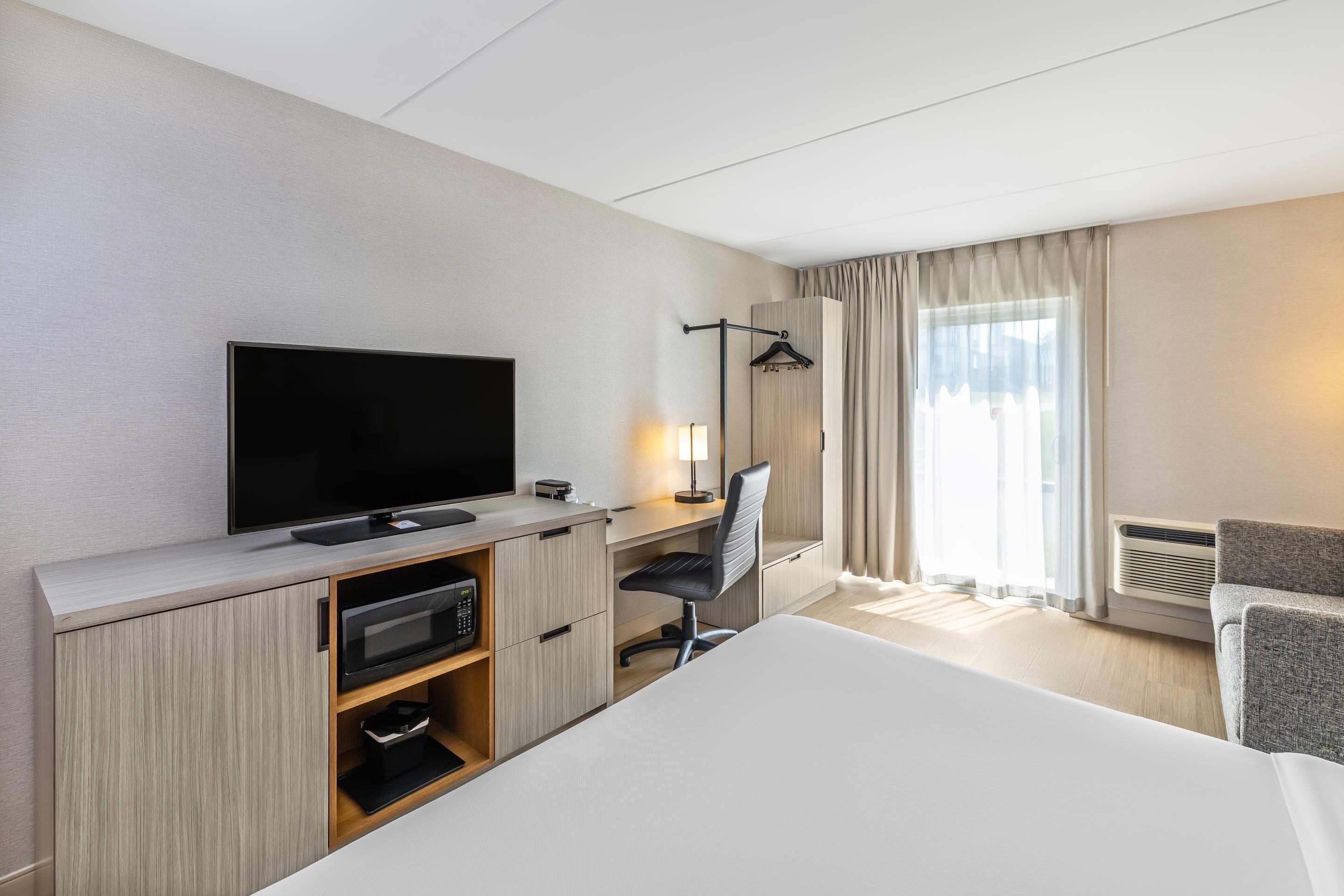 Une chambre d’hôtel équipée d’une télévision à écran plat, d’un bureau avec une chaise, d’un lit, d’une armoire et d’une fenêtre avec des rideaux.