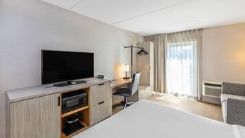 Une chambre d’hôtel équipée d’une télévision à écran plat, d’un bureau avec une chaise, d’un lit, d’une armoire et d’une fenêtre avec des rideaux.
