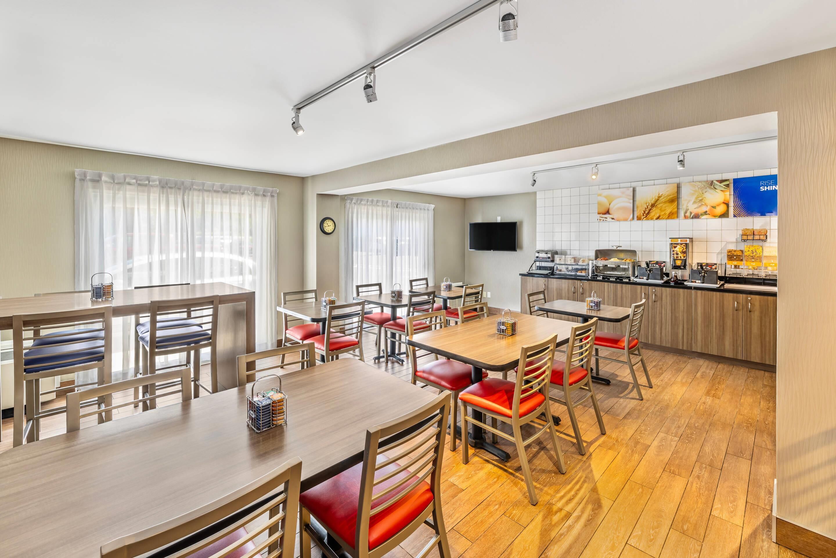 A hotel dining area with multiple tables and chairs, a kitchen area with a flat-screen TV, and a clock on the wall.