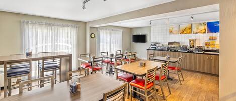 A hotel dining area with multiple tables and chairs, a kitchen area with a flat-screen TV, and a clock on the wall.