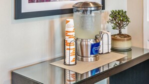 A reception desk with a framed painting, a water dispenser, a potted plant, and stacked cups. - Comfort Inn (Fredericton)