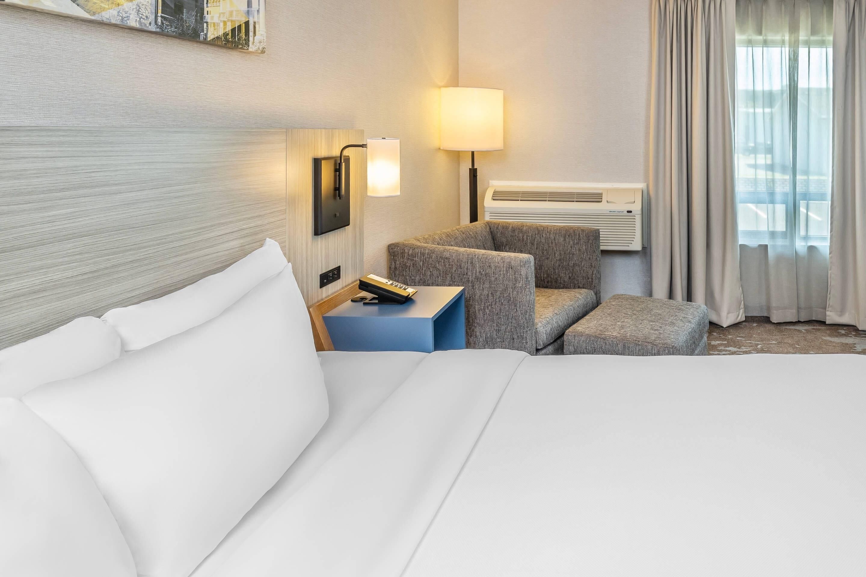Una habitación de hotel con cama, escritorio con lámpara, silla y un pequeño reposapiés.