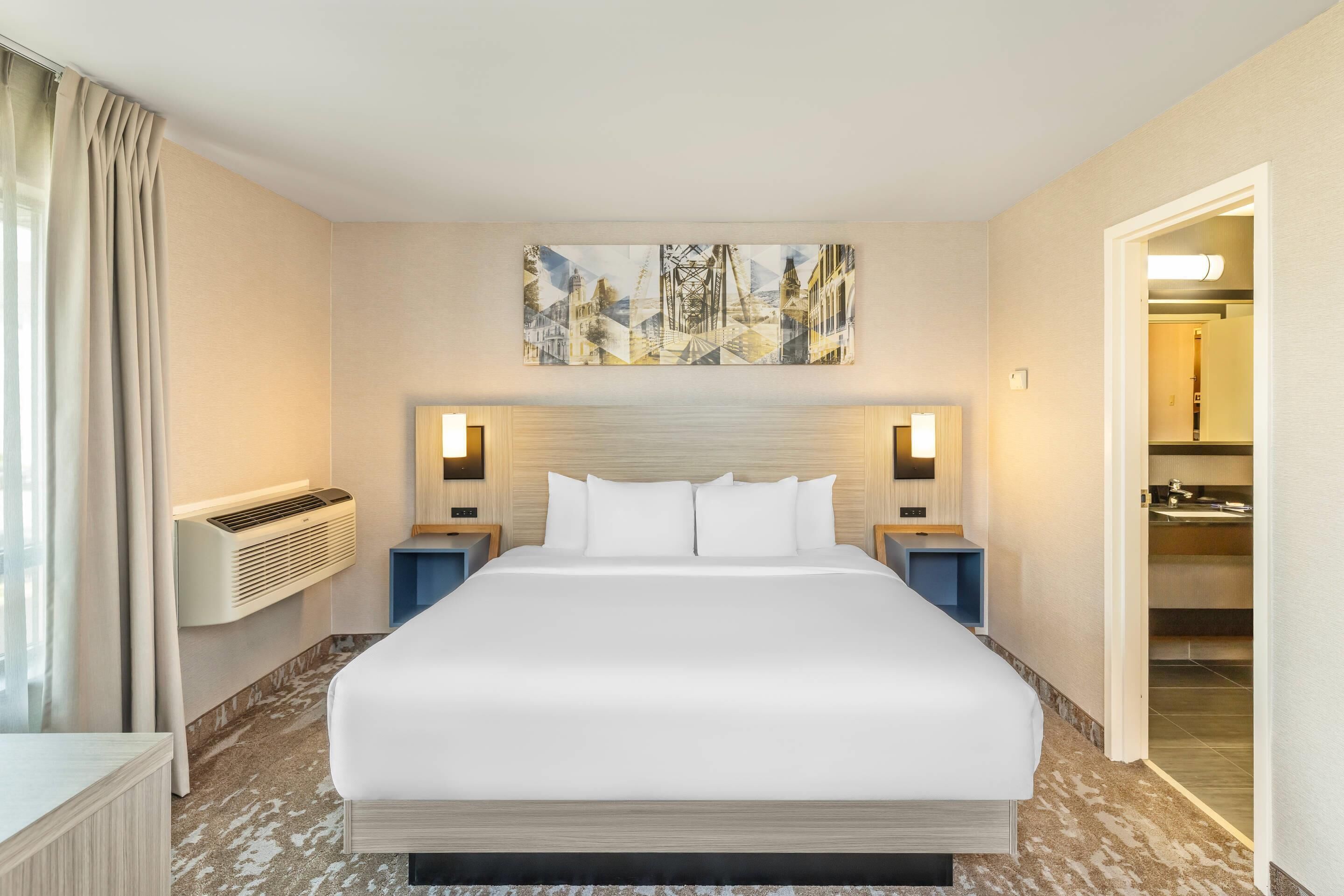 Habitación de hotel con una cama grande, dos mesitas de noche, aire acondicionado de pared, un cuadro sobre la cama y un baño visible a través de una puerta abierta.