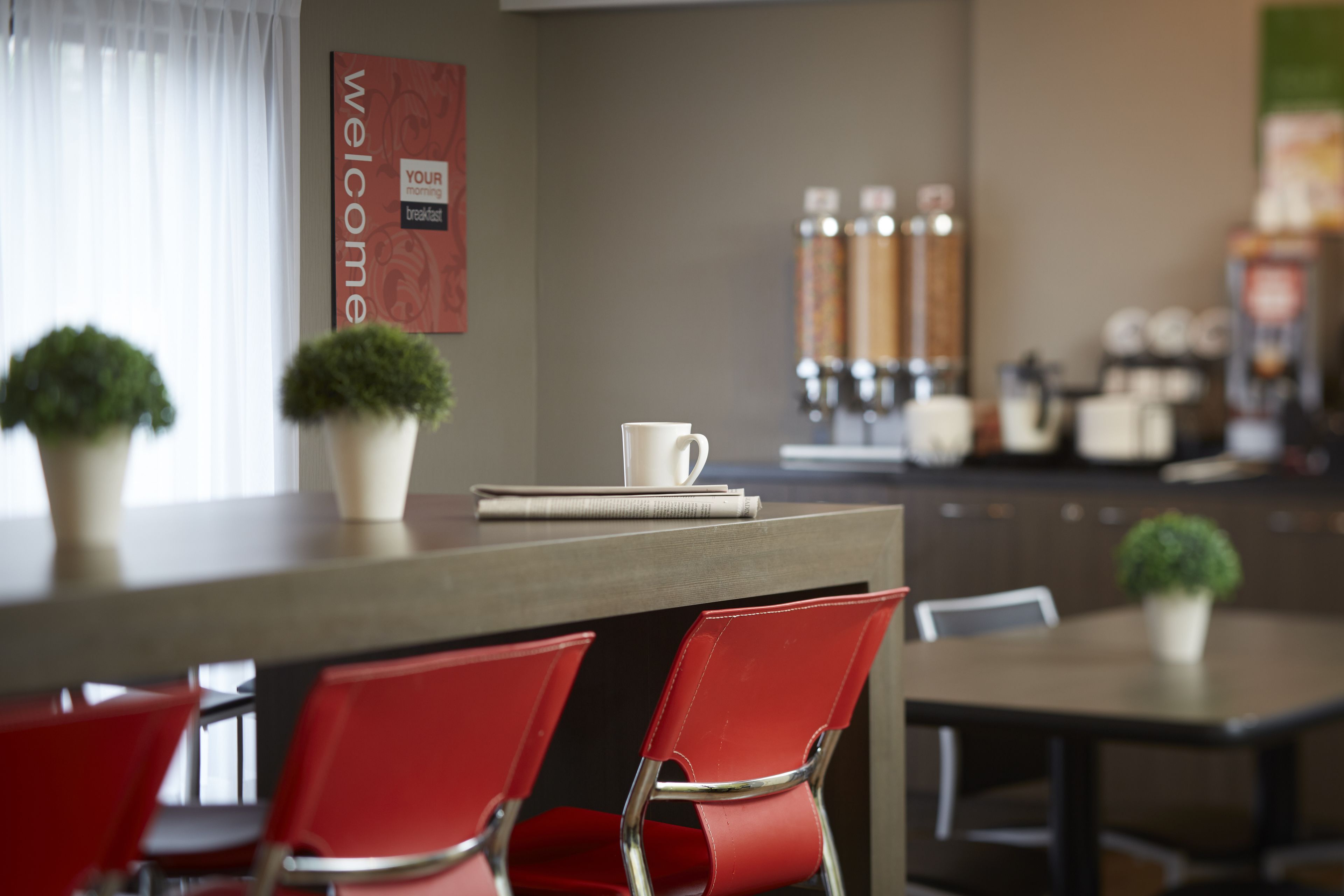 A modern dining area with red chairs, a white coffee cup, and a 'Welcome' sign.