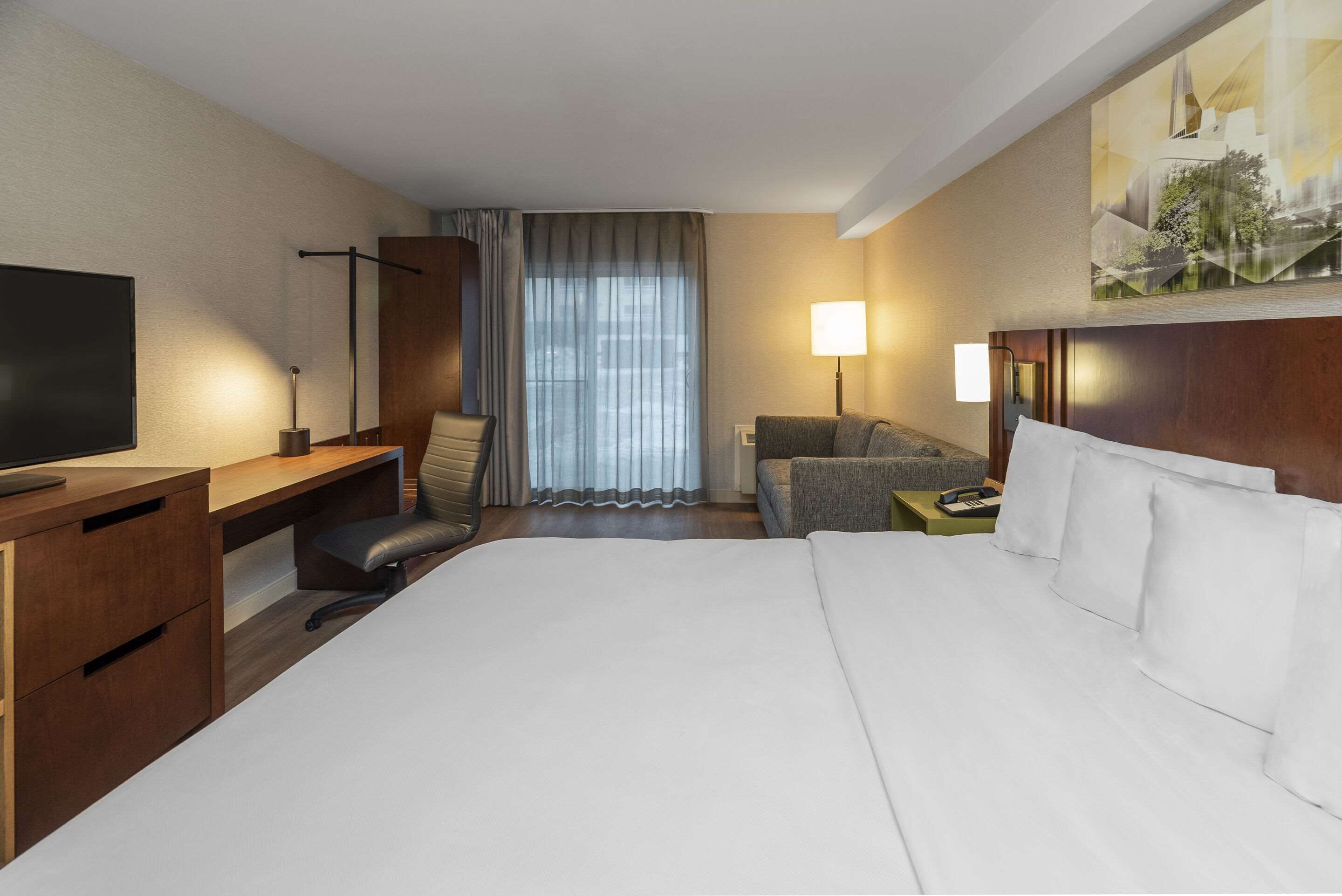 A hotel room with a large bed, a desk with a chair, a sofa, a TV, and a window with curtains.