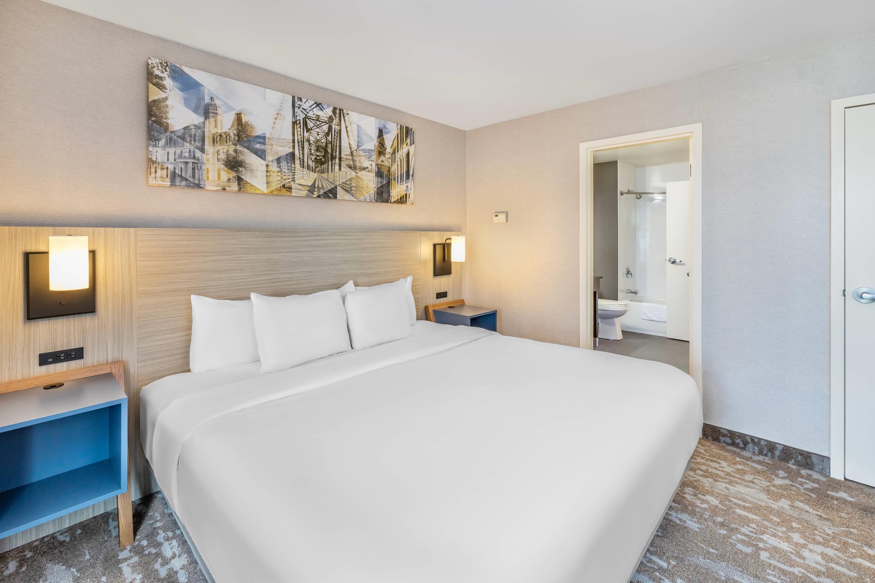 Hotelkamer met een groot bed, nachtkastjes, een kunstwerk aan de muur en een badkamer zichtbaar door een geopende deur.