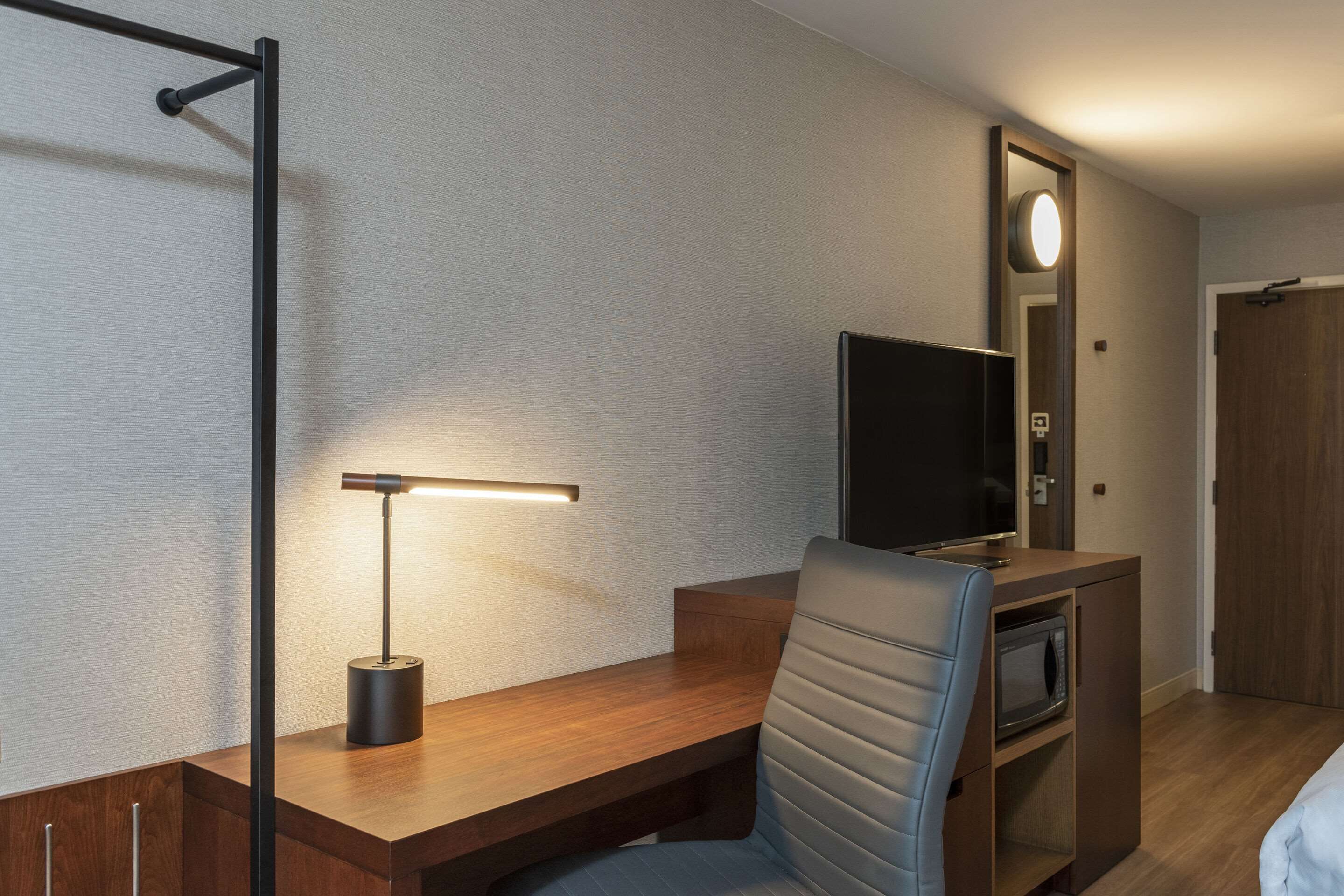 A hotel room with a wooden desk, a chair, a flat-screen TV, a microwave, and a mirror with a light.