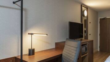 A hotel room with a wooden desk, a chair, a flat-screen TV, a microwave, and a mirror with a light.