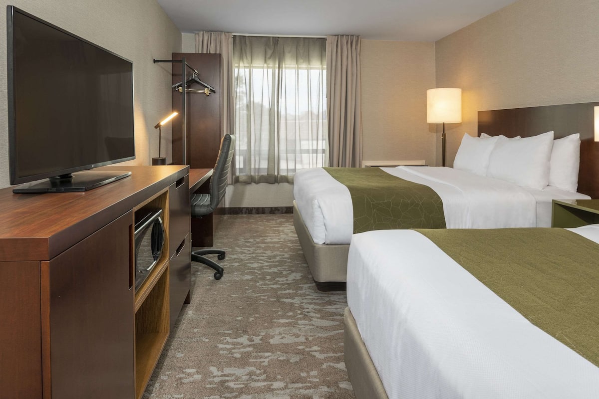 A hotel room with a flat-screen TV, a desk with a chair, two beds, a lamp, and a view of the outdoors through the window.