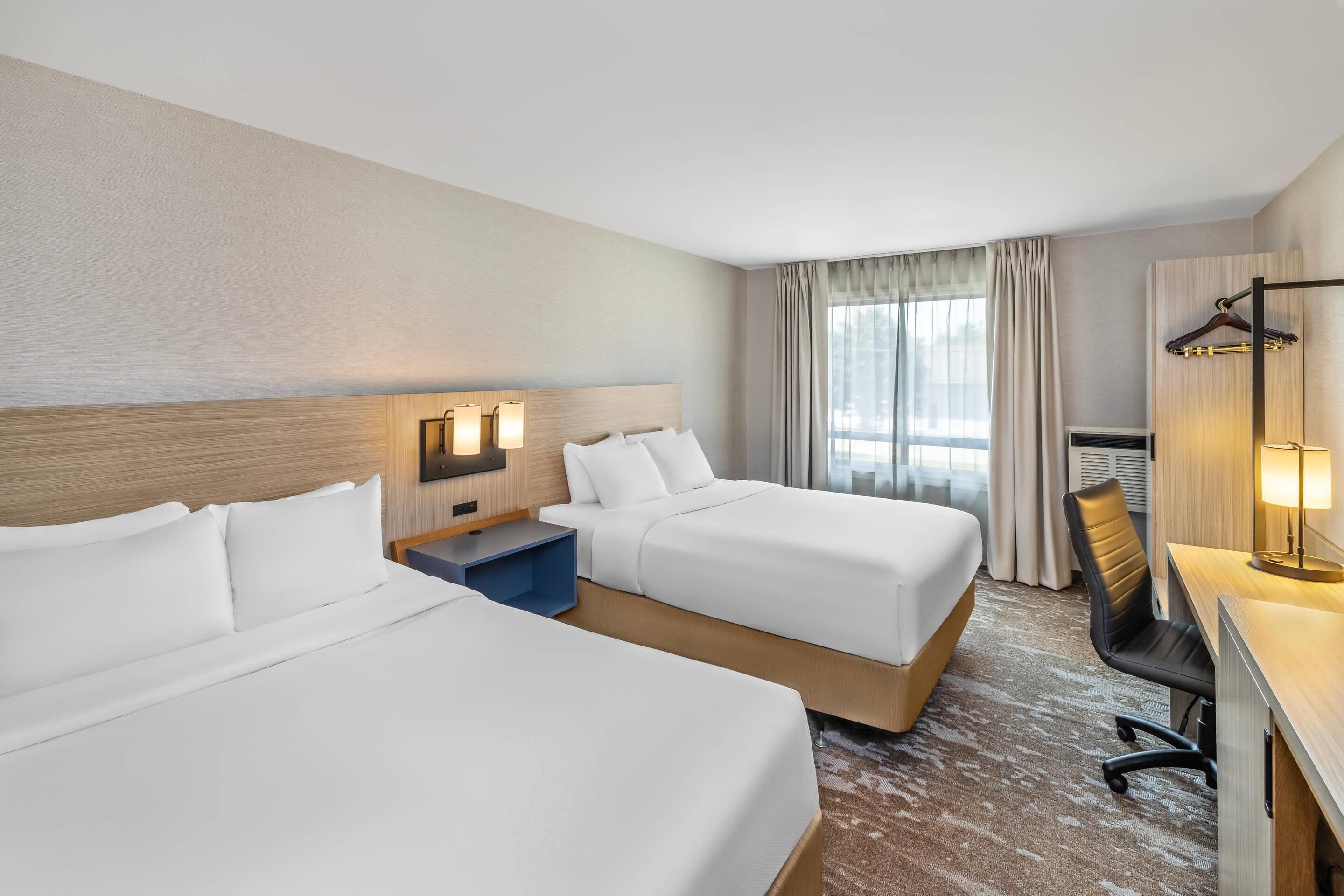 A hotel room with two beds, a desk with a chair, a desk lamp, a TV, and a window with curtains.