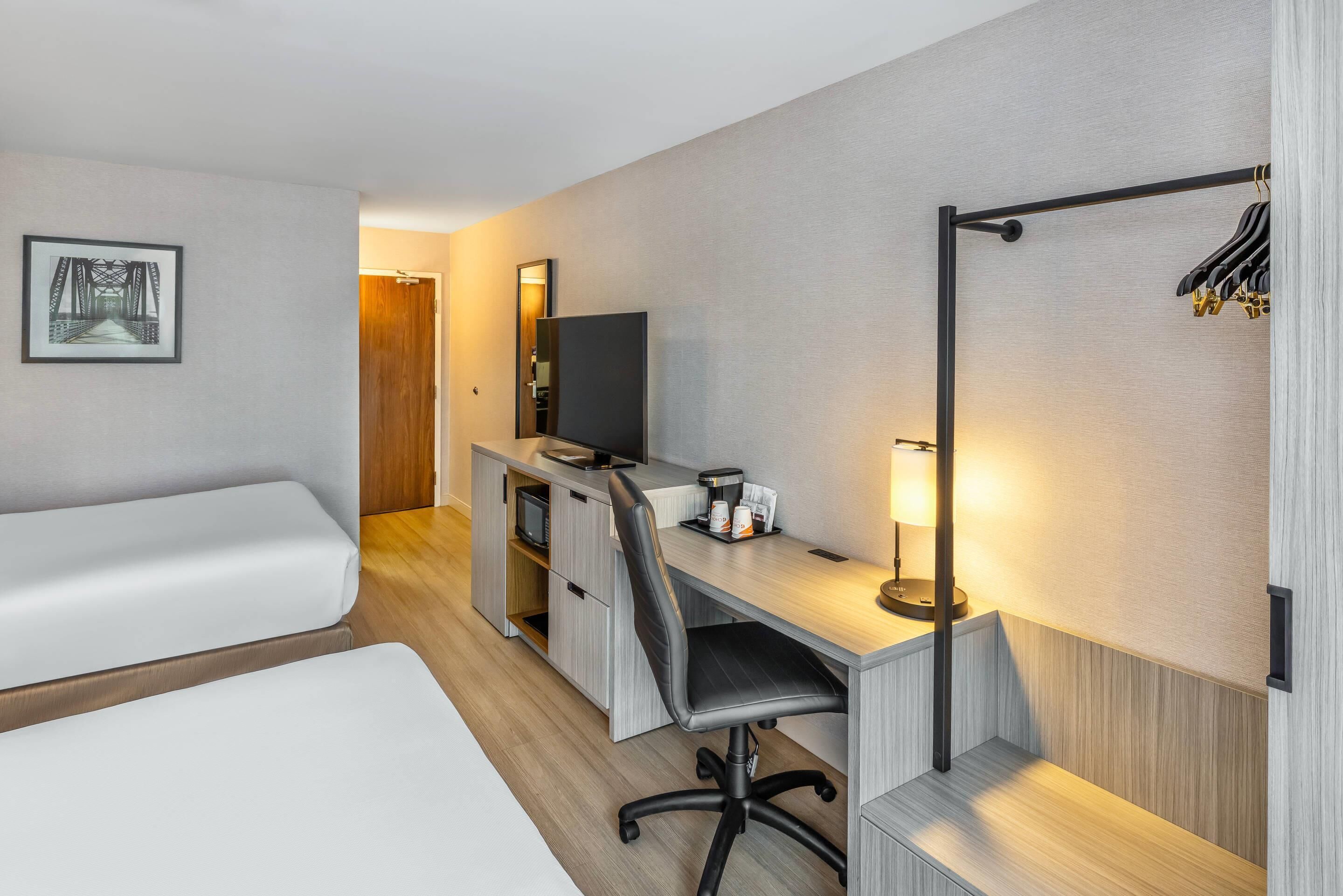 A hotel room with a bed, a desk with a chair, a television, a mirror, and a coat rack.