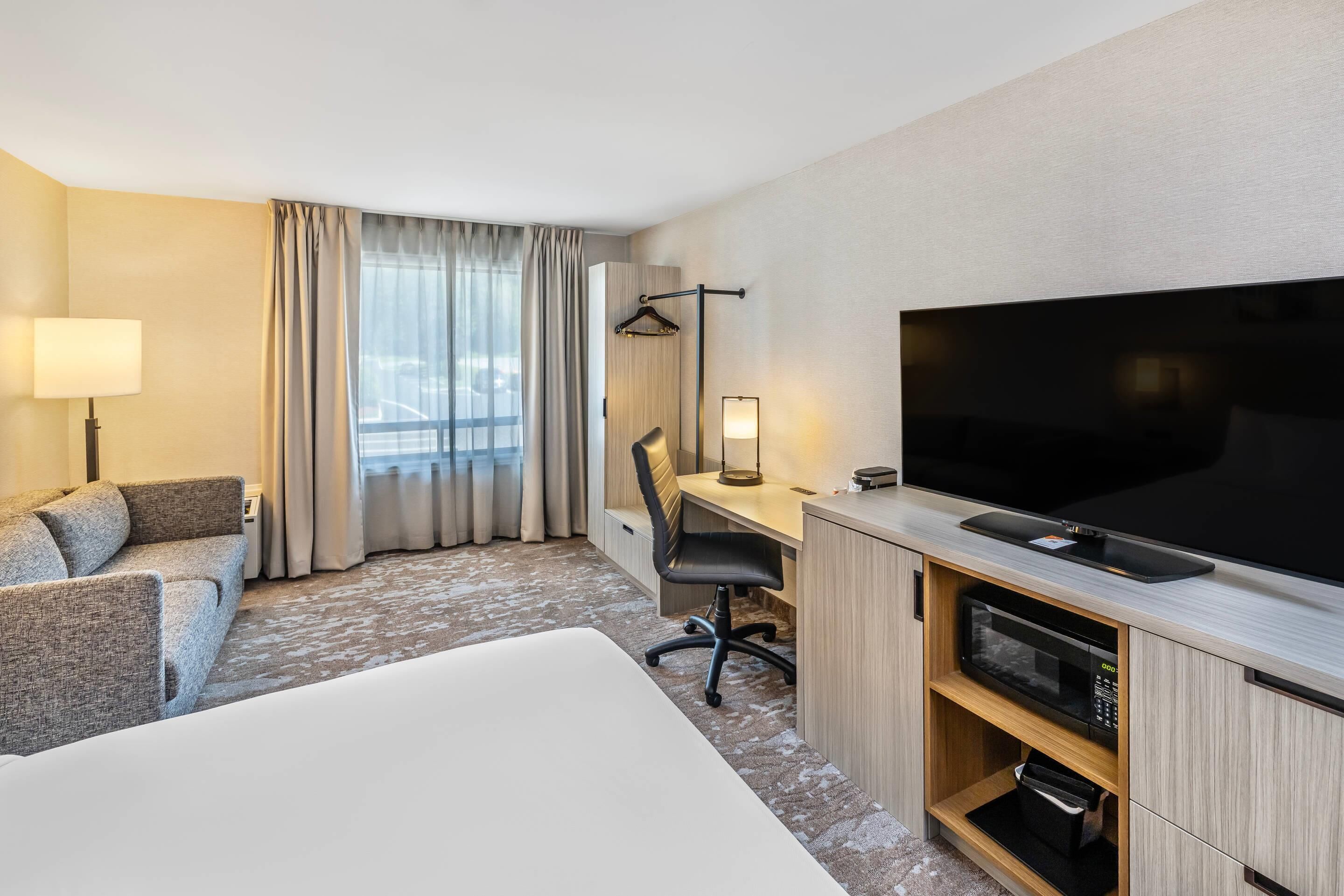 A hotel room with a large bed, a desk with a TV, a chair, a lamp, and a window with curtains.