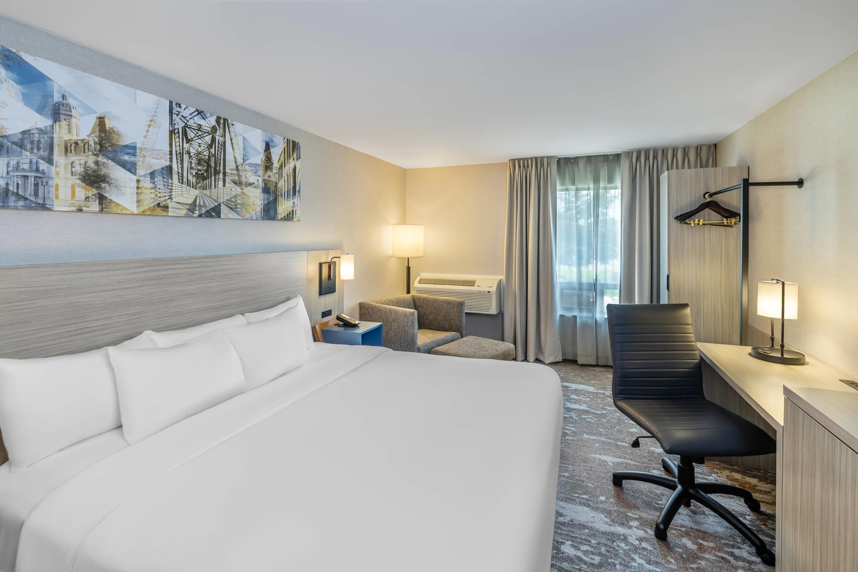 A hotel room with a large bed, a desk with a chair, a small table, a lamp, and a painting on the wall.