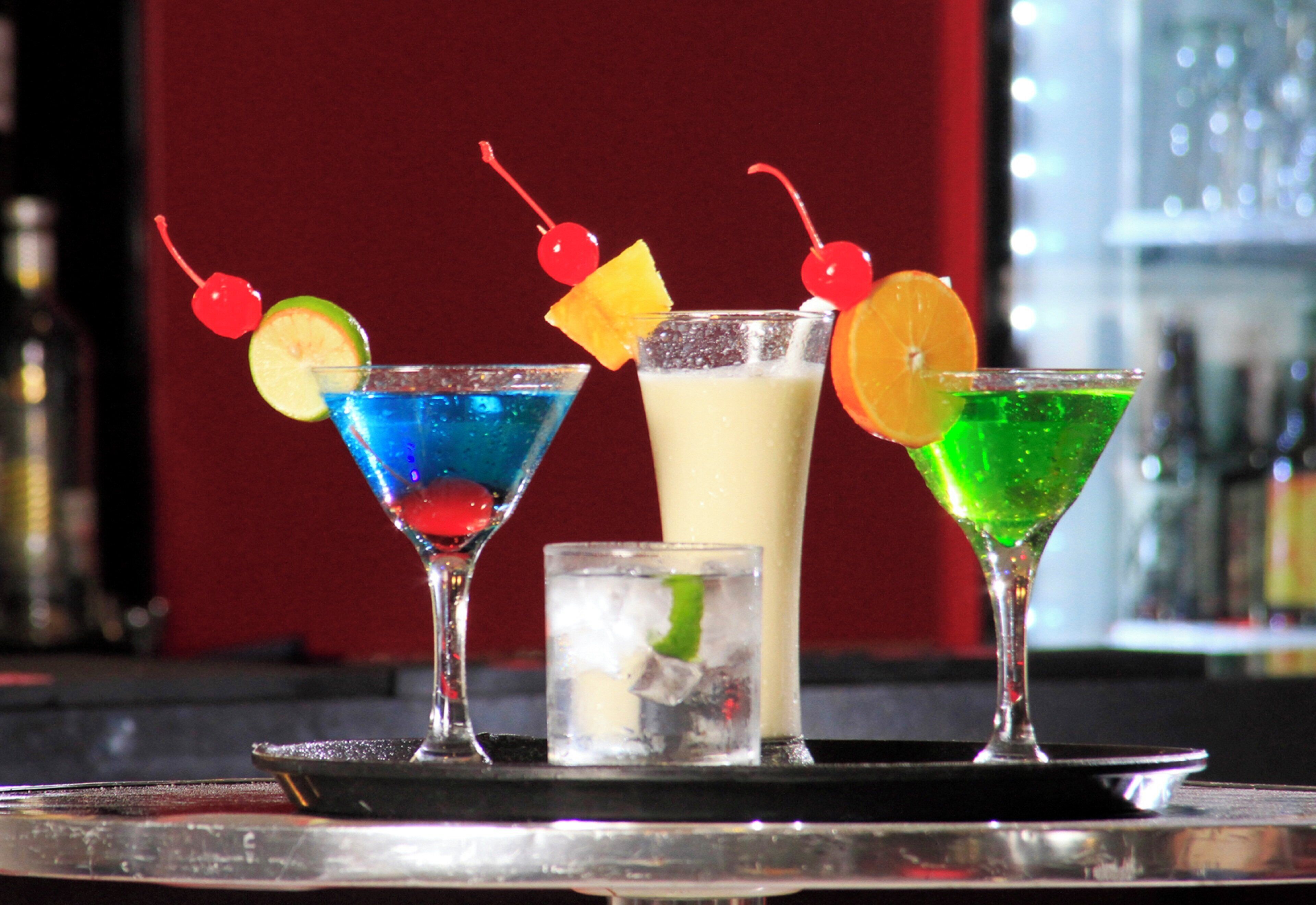 Un bar con tres cócteles diferentes, cada uno decorado con frutas variadas y una cereza, además de un vaso pequeño de agua con una rodaja de lima.