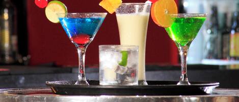 Un bar con tres cócteles diferentes, cada uno decorado con frutas variadas y una cereza, además de un vaso pequeño de agua con una rodaja de lima.